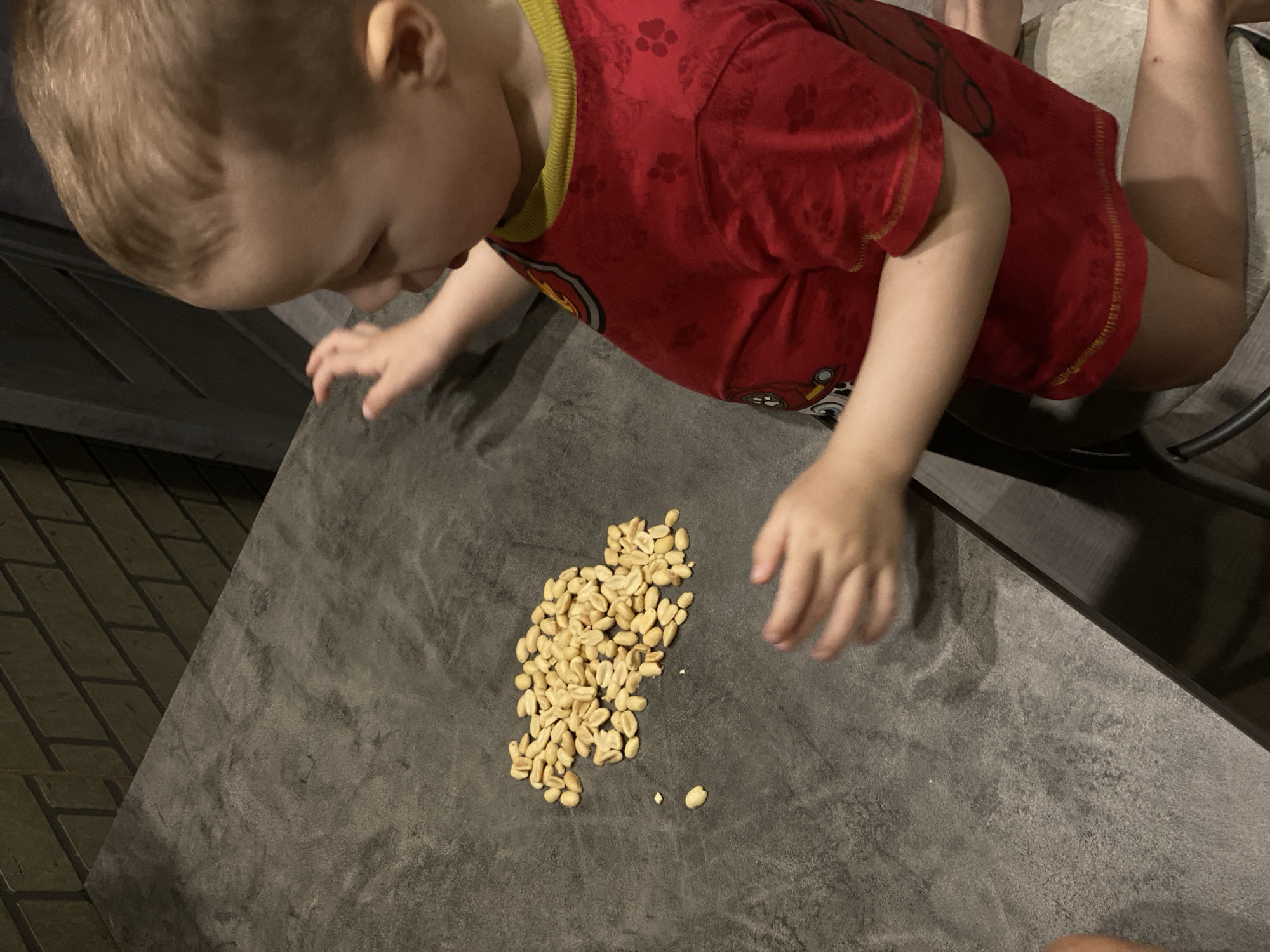 An indoor scene in Sochi, Russia, depicts a young child interacting with a pile of shelled peanuts on a dark gray, textured countertop. The child, visible from the mid-torso upward on the right side of the frame, has light brown hair and fair skin, and wears a red short-sleeved shirt with yellow trim and "Paw Patrol" graphics. The child is observed looking downward towards a cluster of approximately 50-70 unshelled peanuts located slightly left of the image center. The child's right hand, open, hovers above the peanuts, indicating an intent to interact. A dark, brick-patterned wall serves as the background above the countertop. A portion of a dark chair or cabinet is visible to the lower right of the child. The child's left leg, bent at the knee, suggests a kneeling or seated position on a surface not entirely visible within the frame.