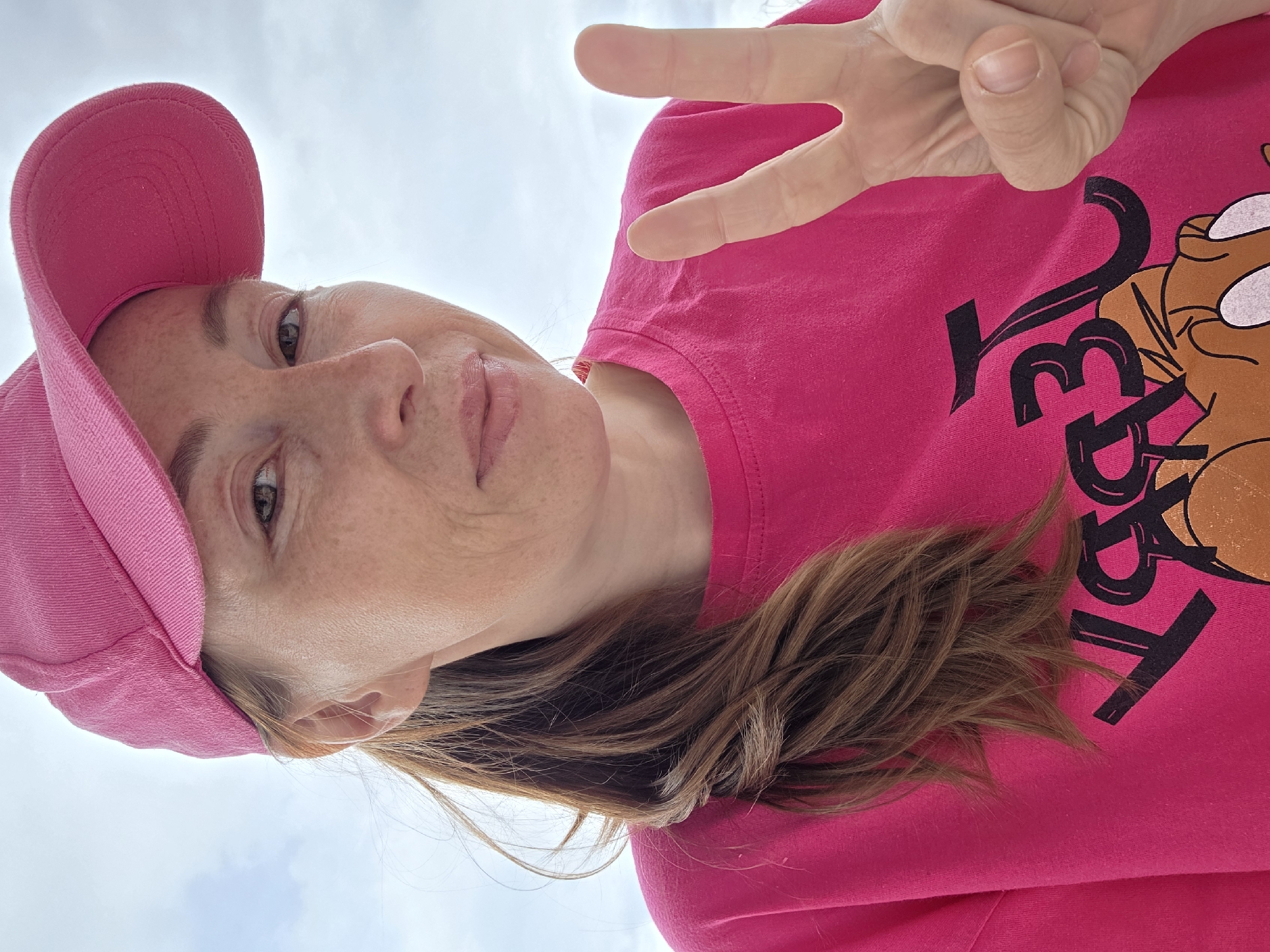 A low-angle selfie captures a person from the chest up, set against an overcast sky. The subject, likely a woman, wears a bright pink baseball cap and a matching pink t-shirt. Her brown hair frames her face. She has light brown eyes and visible freckles on her nose and cheeks, with a subtle smile. Her right hand is raised, displaying a "peace" or "victory" sign. The pink t-shirt features a graphic design: stylized black letters, which appear to spell "VELMA," alongside a brown cartoon dog head with white eyes, strongly resembling Scooby-Doo. The background is a bright, cloudy sky, indicating an outdoor daytime setting. The image conveys a casual and pleasant mood. This media was captured in Chaikovsky, Russia.