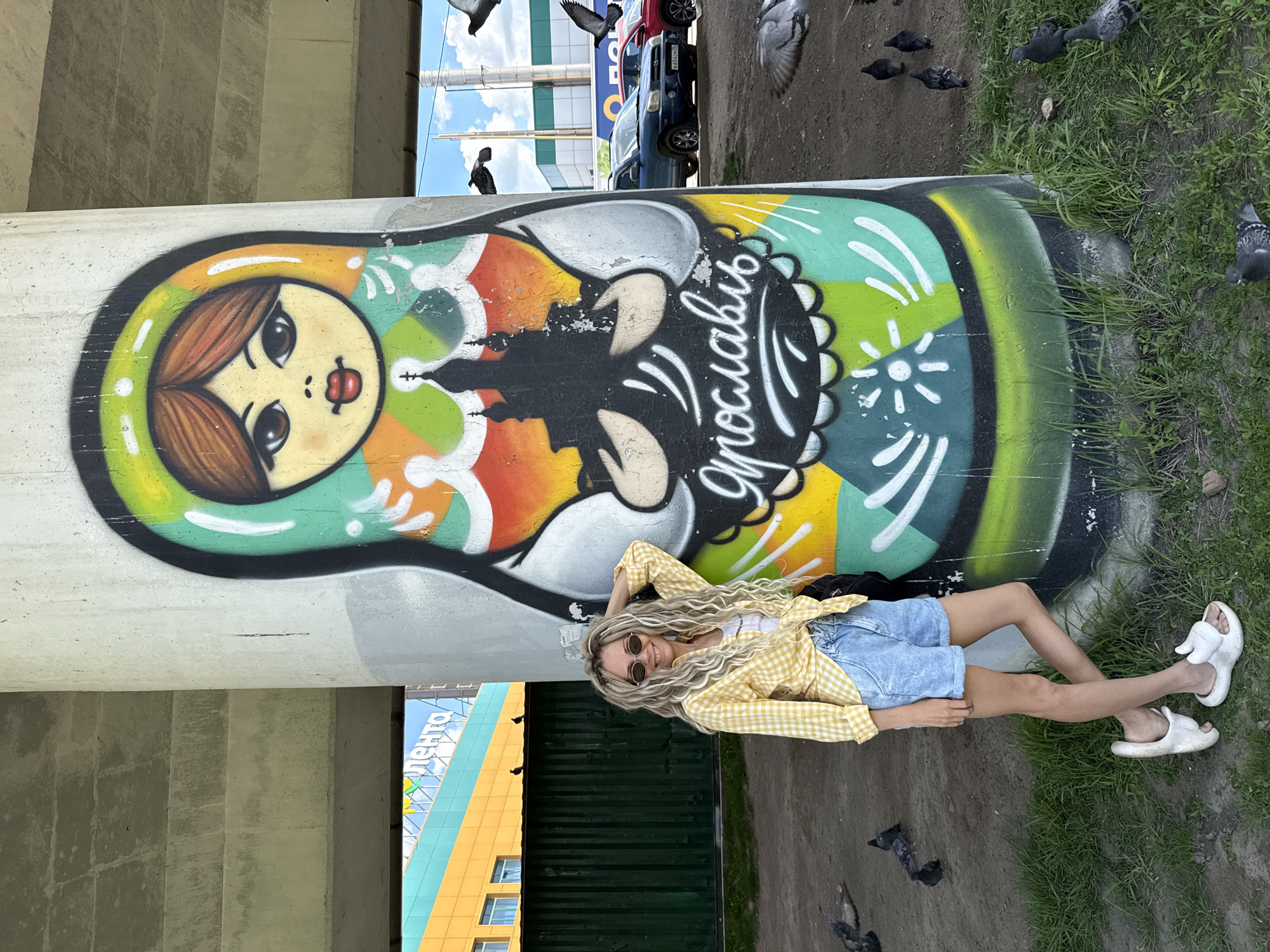 A woman lies on the ground next to a column that features a painting of a matryoshka doll, with pigeons around her.
Location: Заволжье, Russia.
Subject: A woman with blonde dreadlocks, sunglasses, a yellow and white checkered shirt, denim shorts, and white sandals is reclining. She is resting one arm behind her head.
Objects:
- A column with a large, vertical matryoshka doll painting. The doll has a colorful design with Russian script.
- Pigeons are scattered on the ground near the woman and the column.
- Grass and a paved surface surround the area.
- Cars and buildings are visible in the background.
- A black backpack is near the woman.
Actions:
- The woman is lying down, posing for the photo.
- Pigeons are standing and flying in the vicinity.
Interactions: The woman is interacting with her environment by posing near the painted column. Pigeons are moving around in the space.
Environment: The scene appears to be under an overpass or bridge support in a city. The setting includes urban elements like roads, buildings, and a painted column. It appears to be daytime, under natural light.
