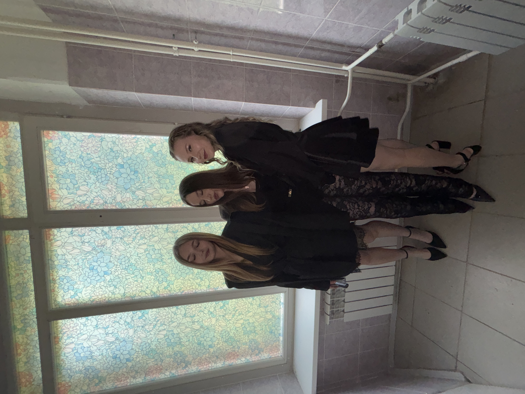 Three young women are posing in front of a large, decorative window in what appears to be a school hallway. The window is comprised of multiple panes, each filled with a stained-glass-like pattern of floral motifs in shades of blue, green, pink, and yellow. The women are dressed in black formal attire. The woman on the left wears a black dress with a lace hem and high heels. The woman in the center wears a black top and lace pants with high heels. The woman on the right wears a black dress with a skirt and high heels. The hallway has tiled walls and floors, with a white radiator visible below the window. Two women have their eyes closed, while the third on the right looks towards the camera. The scene is lit by natural light coming through the window. The location is the Chaikovsky Municipal Pedagogical College in Chaikovsky, Russia.