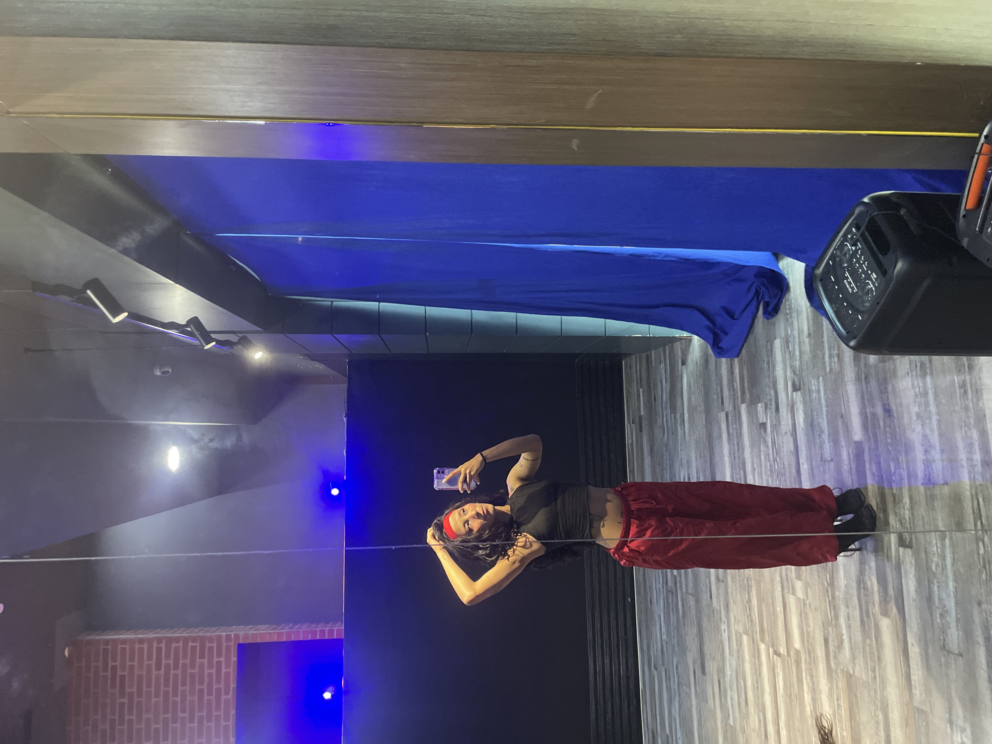 A young woman is standing in what appears to be a dance studio, taking a mirror selfie. She is wearing a black crop top, red satin pants, and black high-heeled boots. She has dark curly hair and a red bandana on her head. She is touching her hair with her right hand and holding a phone in her left hand. The studio has a wooden floor and a dark wall in the background. There are blue lights illuminating the space. In the reflection, a portable speaker is visible on the right side of the image. The location is identified as Pampatar, Venezuela.