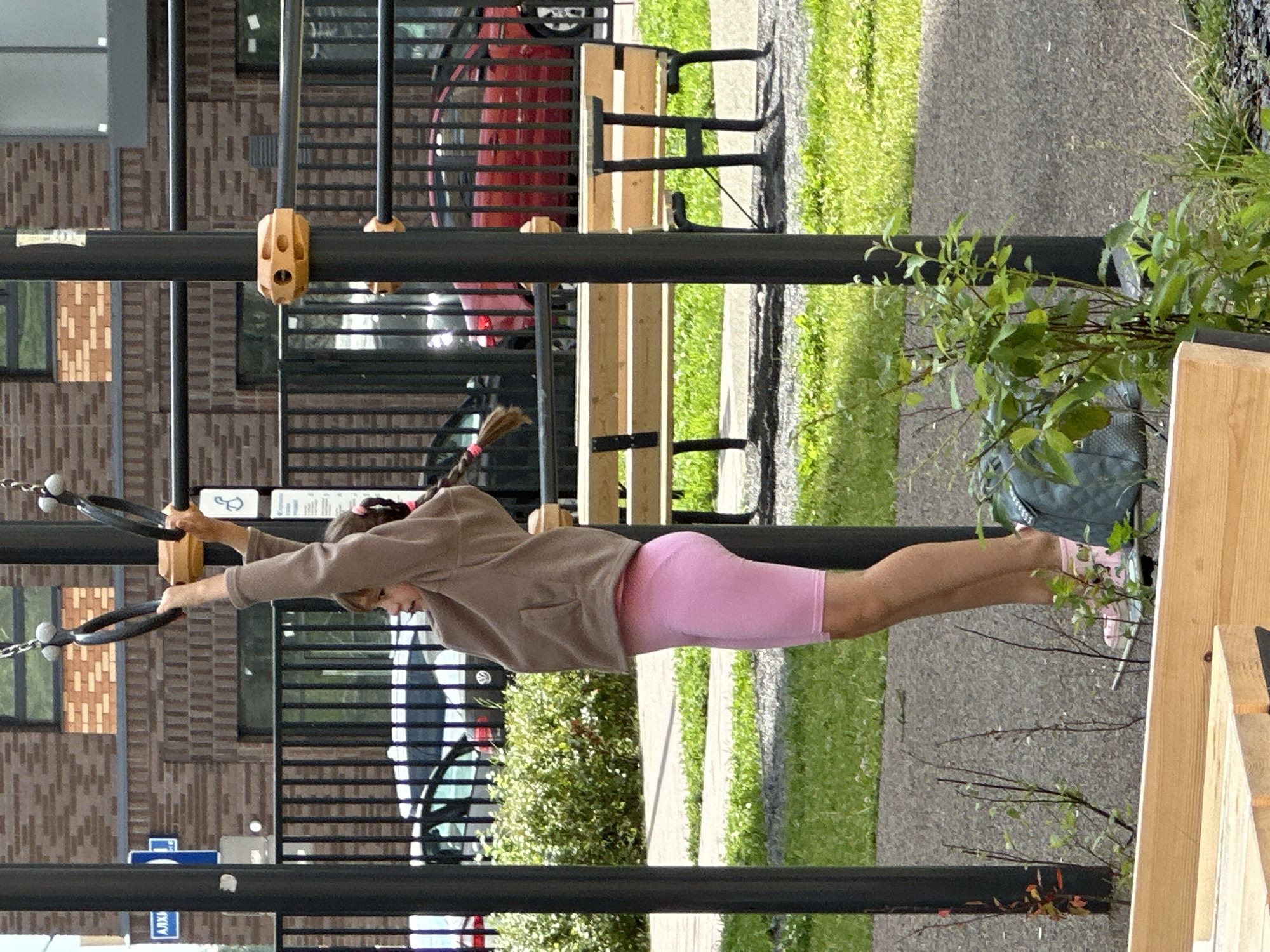 The image shows a young girl with braided hair, wearing a tan hoodie and pink biker shorts, hanging from gymnastics rings in an outdoor playground. She is looking to her left. Behind her, a black metal fence encloses a paved area with a red car parked on the other side. A wooden park bench is visible in the mid-ground. The background features a brick building. The playground is located in Znamya Oktyabrya, Russia.