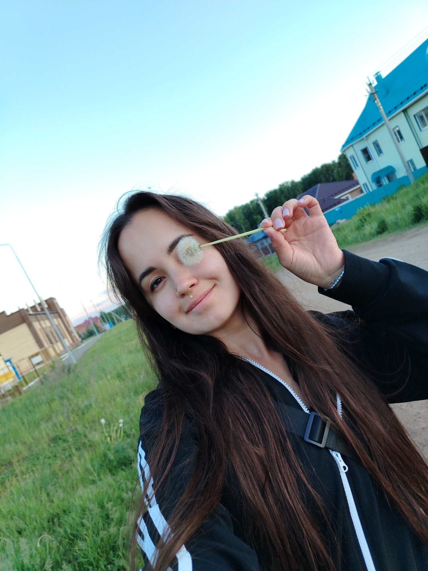 A young woman is featured in a close-up selfie taken outdoors in Noviy, Russia. She has long, dark brown hair and is wearing a black zip-up jacket with white stripes on the sleeves, resembling an Adidas design. A black bag strap is visible across her chest. She has a nose ring on her right nostril and a septum piercing. With her left hand, which has light pink polished nails and a beaded bracelet on the wrist, she holds a dandelion seed head playfully covering her right eye, while smiling softly at the camera.
The scene depicts a clear, bright day, likely late afternoon or early evening, given the soft lighting. The background to her left shows a grassy area with other dandelion puffs, alongside a dirt path and a distant light-colored building. To her right, a prominent two-story house with a distinct bright turquoise roof and light-colored walls stands, with a utility pole nearby. The sky above is a clear, light blue. The overall impression is one of playfulness and contentment. No discernible text is visible.