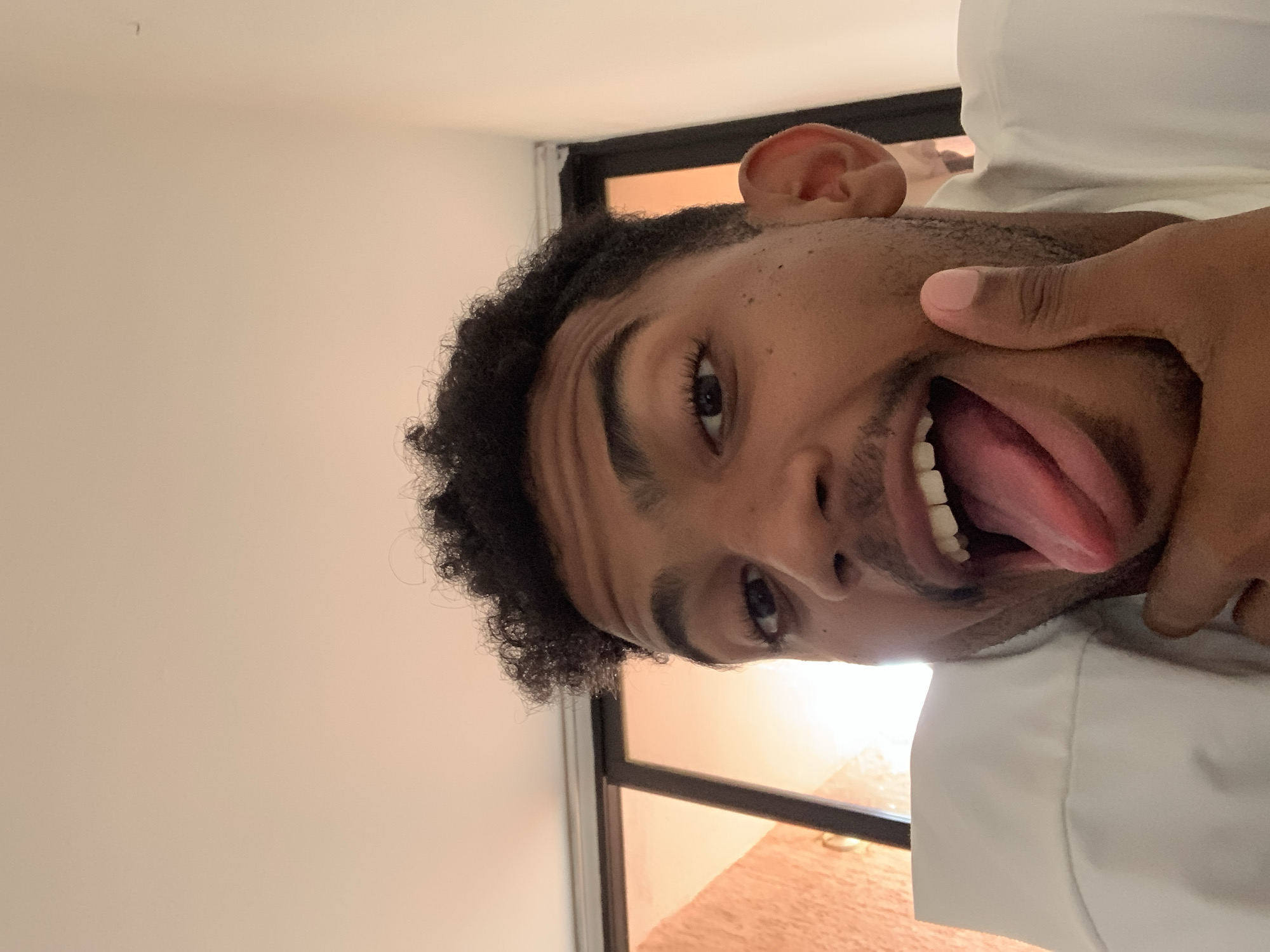A close-up portrait photograph features a person, likely male, with a playful and humorous expression. The person has dark, curly hair, dark eyes, and a light brown skin tone. They are seen sticking their tongue out fully, revealing their teeth, while pressing their thumb against the side of their face, near the jawline. This action slightly distorts the cheek and contributes to the comical expression. The person is wearing a white collared shirt, part of which is visible on the right side of the frame.
The background on the left and top consists of a plain, light-colored wall. Behind the person, a dark window frame is visible, through which warm, soft light streams, suggesting either early morning or late afternoon light. The overall lighting is warm. No specific identifying objects for the location are visible within the image itself, but the context provided for the image is Porlamar, Venezuela. There is no visible text in the photograph.