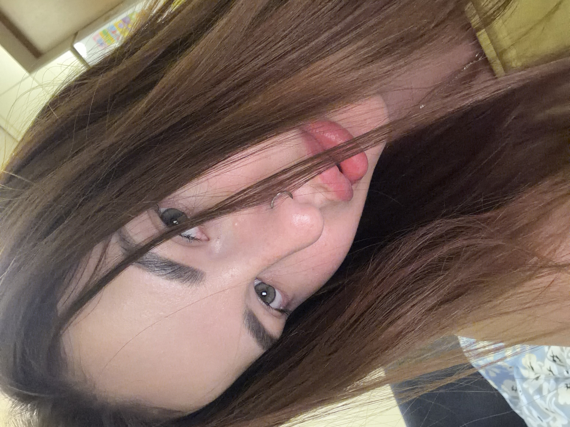 A selfie shot depicts a young woman with brown hair and visible fair skin, seemingly taken indoors. Her eyes are visible and appear to be grey-green. Her tongue is protruding slightly. The picture is at an angle, and part of what looks like clothing with a flower pattern is visible on the right side of the frame. The location is not discernible from the image. There is no indication of Krasnodar, Russia or any specific location.
