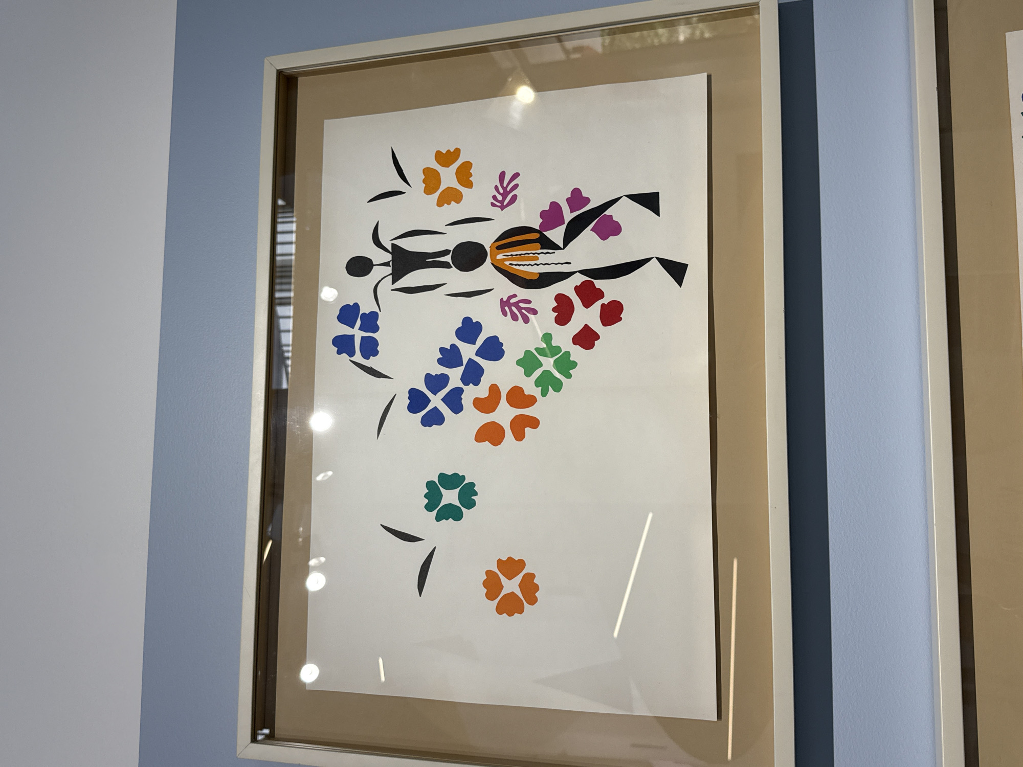 A framed artwork is displayed on a light blue wall. The artwork features a white background with black, red, orange, green, blue, and purple cutouts. The cutouts form abstract shapes, including flowers and two stylized human figures. The figures appear to be stacked or interacting in some way, with one figure balanced on top of the other. The overall impression is that of a collage or paper cutout composition. The framing is a light color, possibly white or cream, with a glass overlay. The lighting in the room creates reflections on the glass. The image does not provide enough information to determine the specific location in Moscow, Russia.