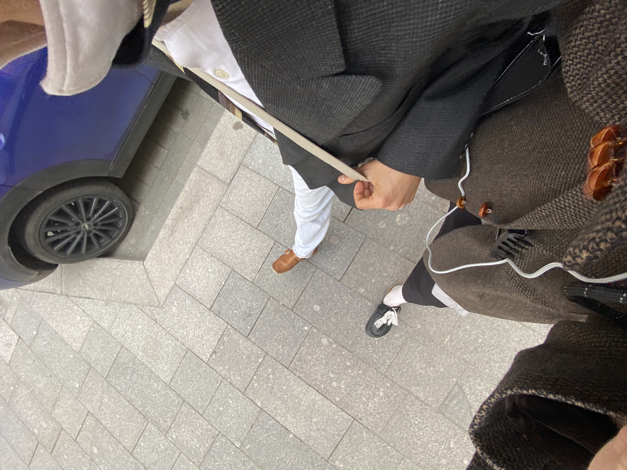 From an elevated perspective, the image depicts a street scene adjacent to a blue vehicle, possibly in Moscow, Russia, evidenced by the architecture and style of clothing. Two individuals are present, both partially visible. One individual is wearing a dark gray coat over a white shirt and white pants paired with brown leather shoes. The other is wearing a brown tweed-like coat over a black lower garment, white socks, and black shoes with white laces. The environment consists of gray tiled pavement.