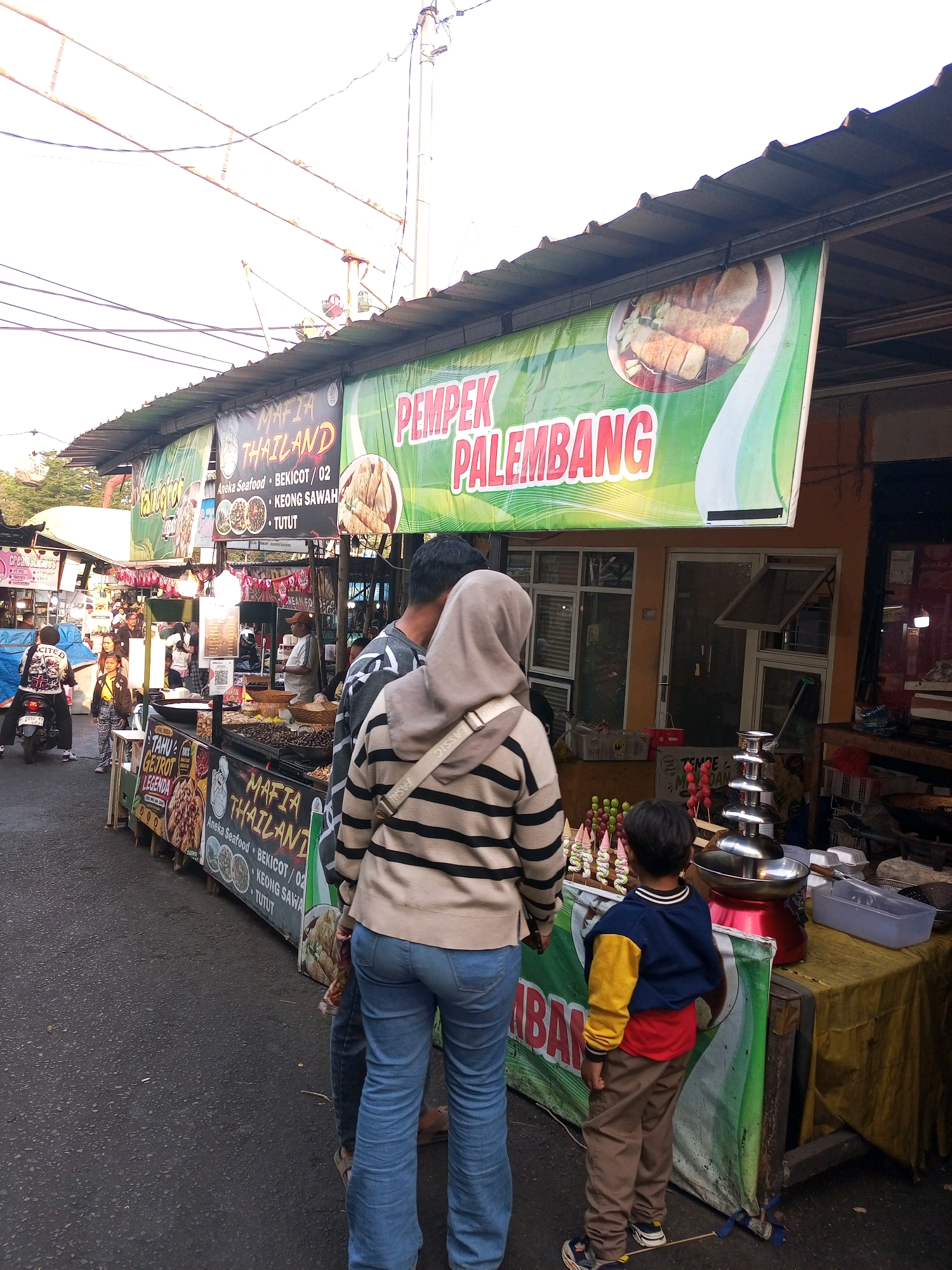The image depicts an outdoor food stall scene in the city of Batu, Indonesia, likely at night or during early evening due to the lighting. The focus is on a long food stall displaying various food items and banners. A prominent banner reads "PEMPEK PALEMBANG" in large red letters, indicating a specialty dish. Another banner features "MAFIA THAILAND" with text like "Aneka Seafood," "Bekicot/02," "Keong Sawah," and "Tutut."
In the foreground, two individuals are partially visible. One is a person wearing a beige and black striped sweater and blue jeans, facing away from the camera. Another person, a young boy in a blue and yellow jacket and brown pants, stands next to them, also facing the stall.
The stall itself is laden with food. There's a chocolate fountain with cascading chocolate and a selection of colorful candies on sticks. Various food trays and containers are visible on the counter. Behind the stall, a building with windows and an open window is present.
In the background, other stalls and people can be seen, suggesting a busy market or food street. A motorcycle with a rider is also visible on the left side of the image. The environment appears to be a street with asphalt paving. Overhead, electrical wires and poles are visible against a bright sky, suggesting an urban setting. The overall impression is a vibrant street food scene.