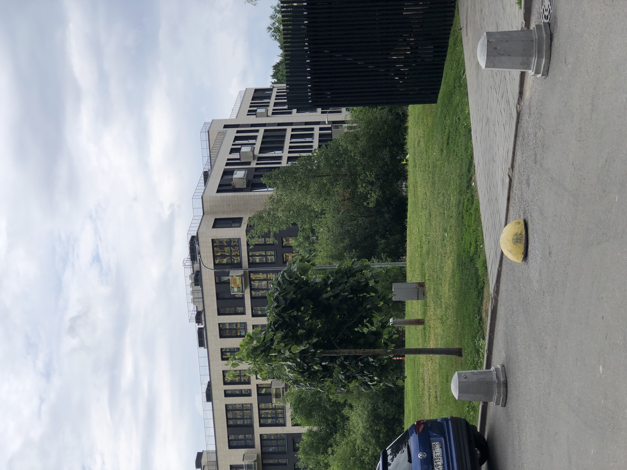 The image shows a modern apartment building with a light-colored facade and dark framed windows. In the foreground, there is a blue car parked on the left, with a license plate visible. A young tree stands in the middle of a grassy area, with a paved walkway to the right. Several concrete bollards are placed along the edge of the road and walkway. A black vertical fence is also visible on the right side of the image. The sky is overcast with gray clouds. The setting appears to be an urban residential area.