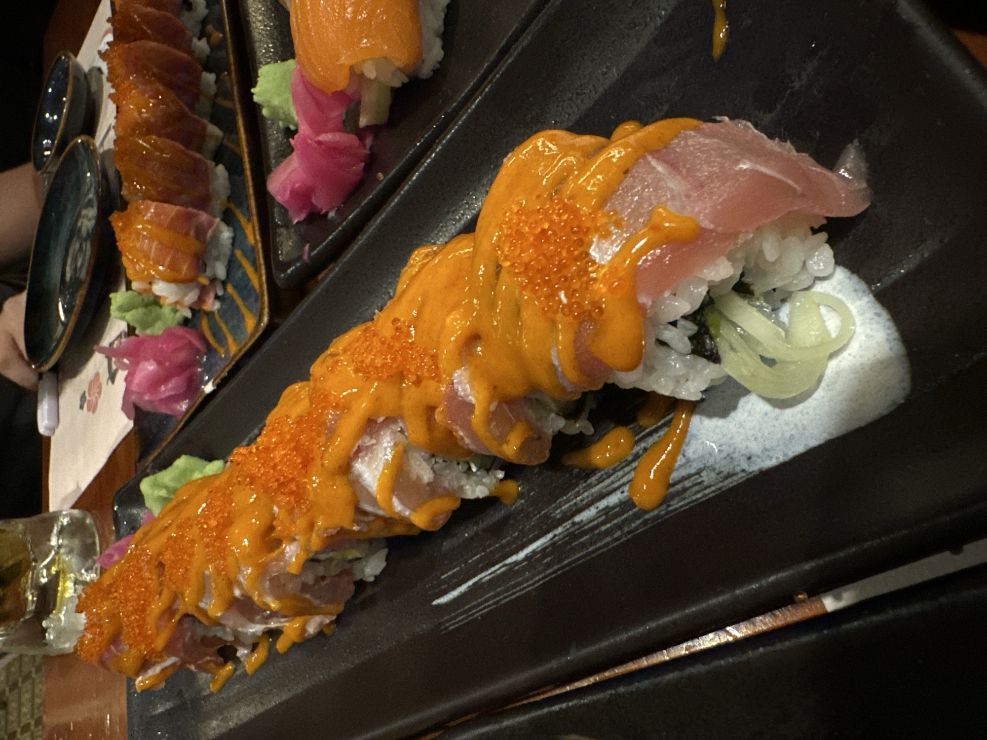 The image displays a close-up, angled shot of an elaborate sushi platter served in a dimly lit restaurant setting, likely in Nha Trang Ward, Vietnam. The foreground features a long sushi roll presented on a rectangular black plate with subtle white and blue decorative patterns. This roll is lavishly covered with a vibrant orange sauce and topped with numerous small, iridescent orange roe, possibly tobiko or masago. Slices of light pink fish, resembling tuna or hamachi, are draped over the individual pieces of sushi, visible beneath the generous sauce. Green, thinly sliced garnishes, possibly spring onions, are also integrated into the roll, and a white, creamy sauce or puree is artfully dabbed on the plate.
In the upper left background, another sushi roll, likely featuring salmon, is partially visible on a different rectangular plate adorned with a distinctive blue and yellow geometric pattern. Beside these rolls are a heap of bright pink pickled ginger (gari) and a small dollop of green wasabi.
To the left, two small, dark blue ceramic dishes, possibly for soy sauce, are visible, along with a clear glass containing ice, suggesting a drink is being served. A human hand or arm is partially discernable in the bottom left, indicating diners are present. The lighting is low and atmospheric, suggesting an evening meal. The presentation of the sushi is detailed and colorful, highlighting fresh ingredients. No text is visible in the image.