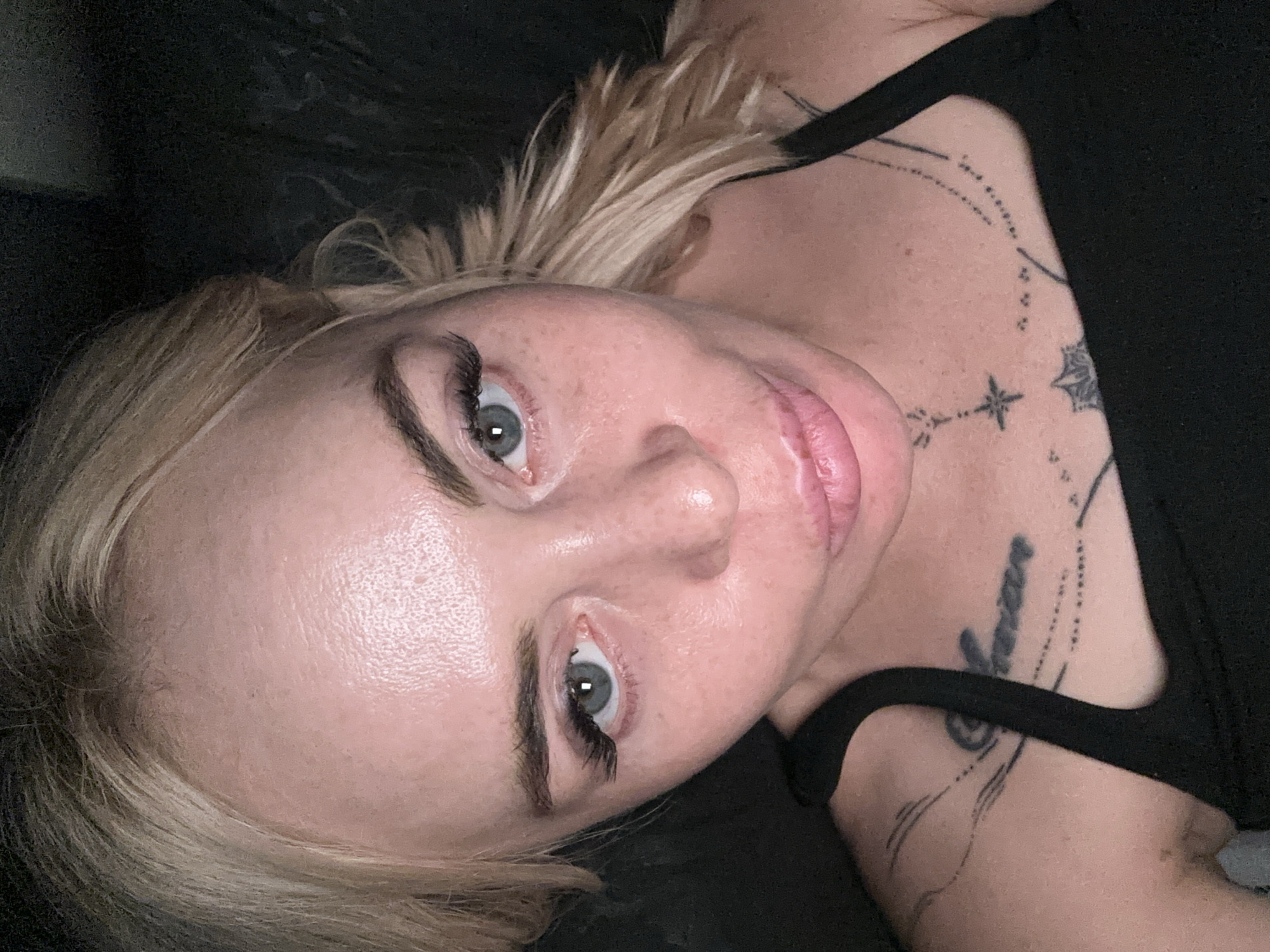 A close-up, overhead shot shows a woman with blonde hair and blue eyes looking directly at the camera. She is wearing a black tank top and has tattoos on her chest and shoulder. The background is dark and blurred.