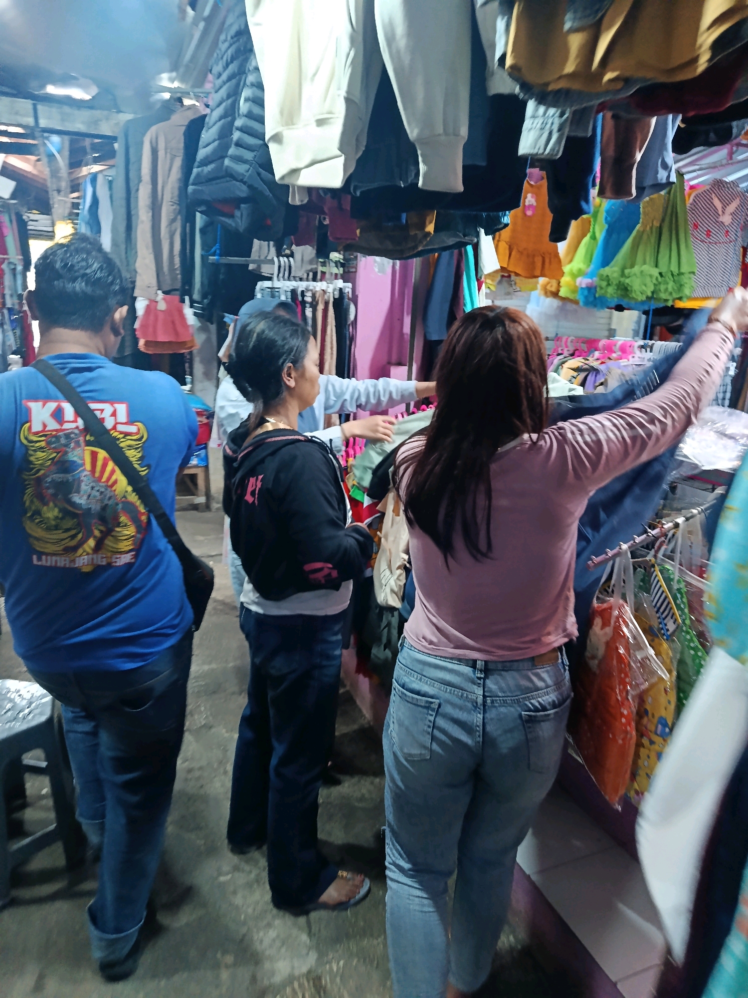 In Senduro, Indonesia, an outdoor market stall is depicted with various clothing items displayed. A man in a blue t-shirt with a black strap across his chest is in the foreground on the left, facing away from the camera. To his right, a woman in a black hooded jacket and jeans is also facing away, looking at the merchandise. Another woman, with long brown hair, is on the far right, wearing a pink long-sleeved shirt and jeans, holding up a piece of clothing. The stall features a variety of clothing, including jackets, shirts, and dresses, hanging from racks and displayed on shelves. The ground appears to be concrete.