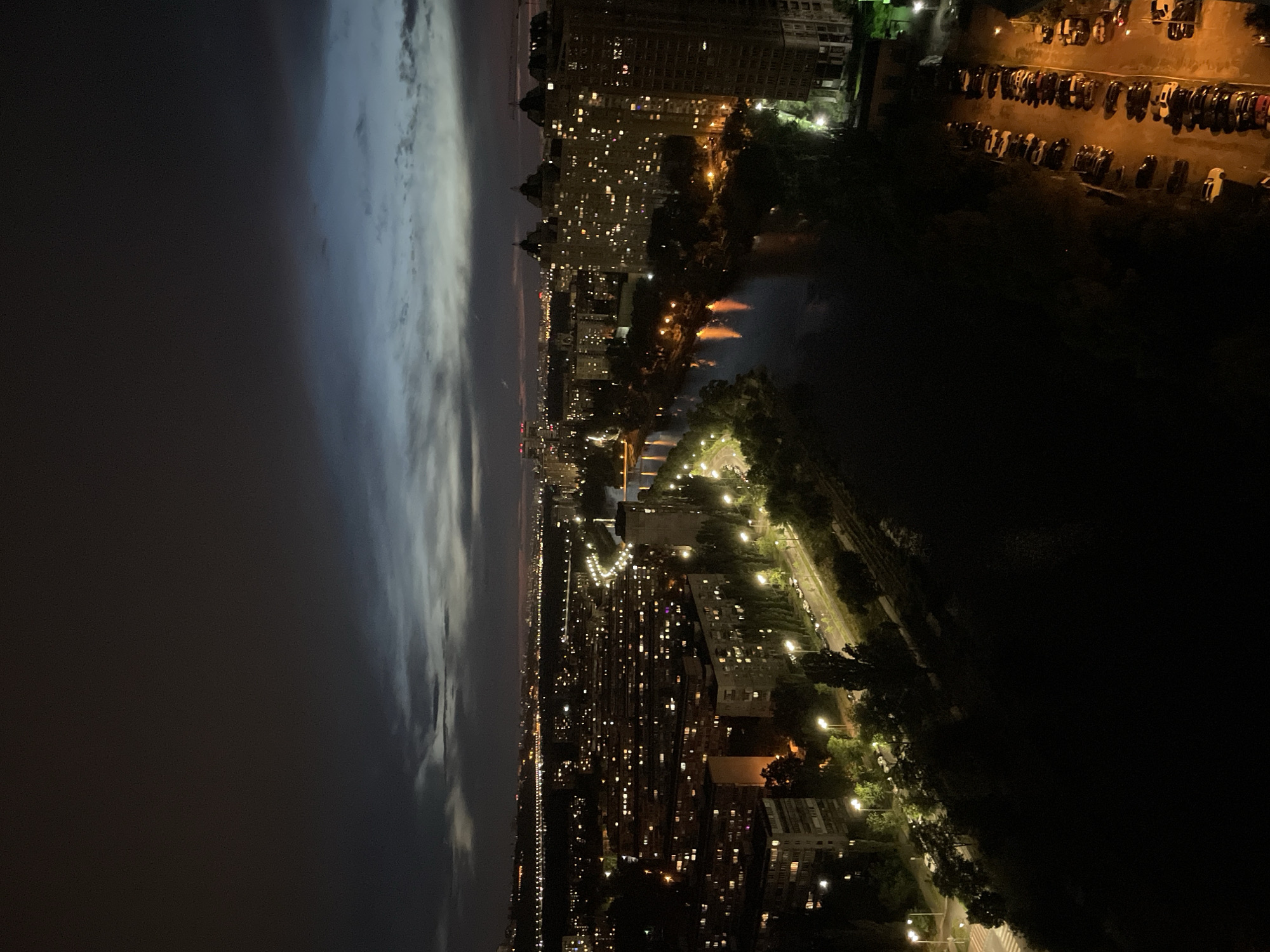 This is a nighttime aerial view of a densely populated urban area in Kyiv, Ukraine, likely taken from a high vantage point. The scene is dominated by the warm glow of city lights reflecting on a body of water, likely the Dnipro River or a canal, which bisects the cityscape. Numerous apartment buildings are visible, with many windows illuminated, indicating occupied residences. Streetlights line roads and pathways, creating a network of light within the city. Some of the buildings appear to be of Soviet-era architecture, characterized by their uniform size and layout. In the foreground, on the right side of the image, a large parking lot is filled with numerous cars. The sky above is dark, with some clouds visible, and a faint horizon line with distant lights suggests the expanse of the city. The overall atmosphere is one of urban activity continuing into the night.