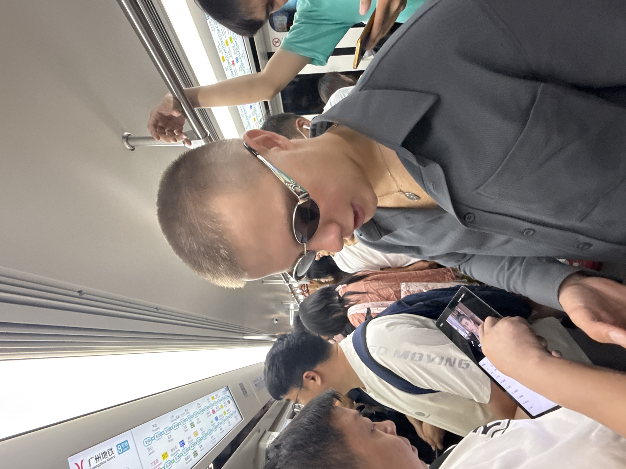 The image shows a crowded subway car in Haizhu District, China. A young man with short hair and sunglasses is the primary subject, facing downwards as if looking at a phone or tablet. He is wearing a dark grey button-up shirt and a necklace with a pendant. Other passengers are visible around him, some holding onto overhead bars or straps. One person in the foreground on the left is holding up a tablet, displaying an image of a person. The subway car interior is illuminated by overhead lights, and a route map is visible on the wall. The atmosphere appears to be that of a typical, busy urban commute.