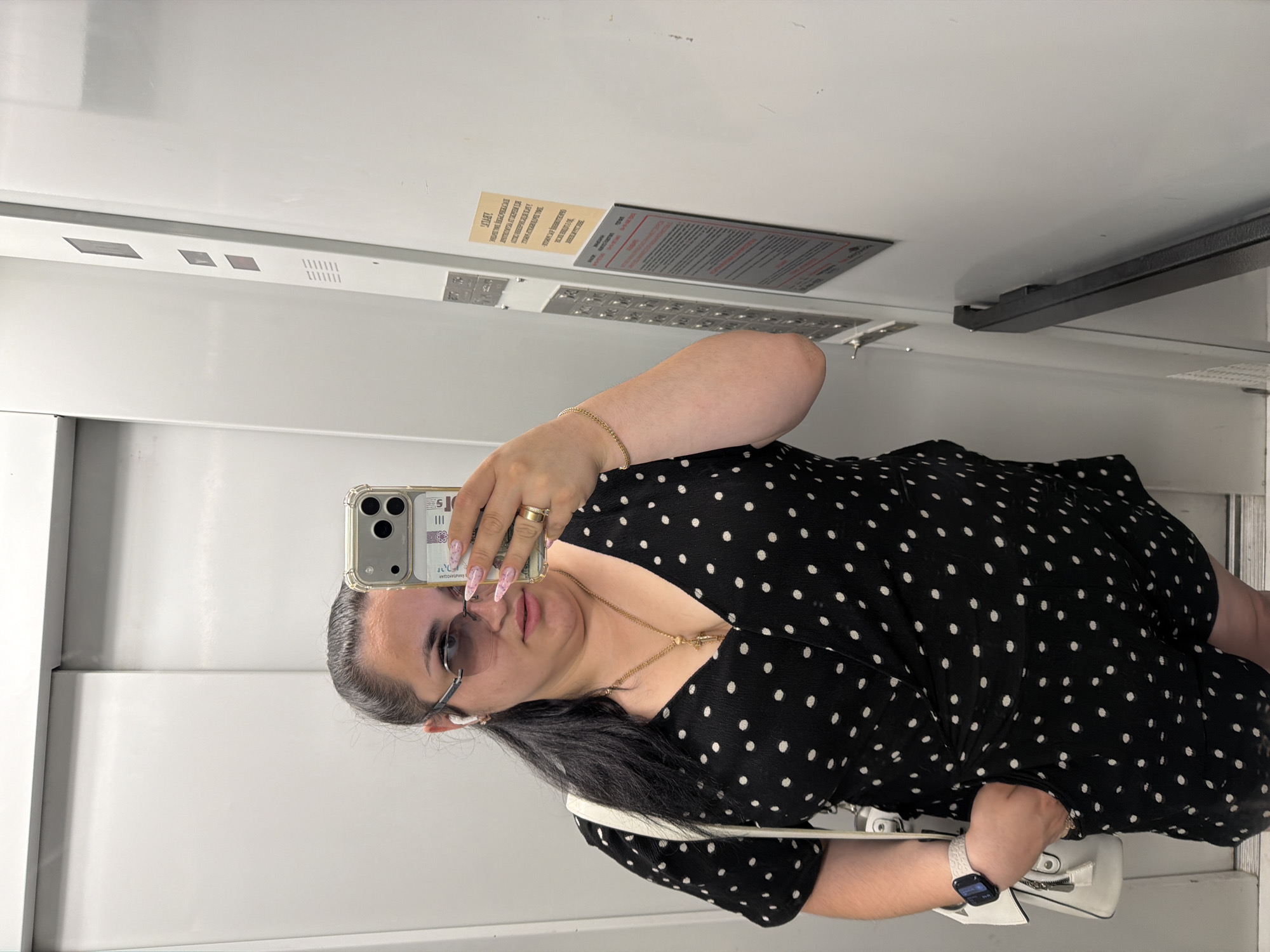 The image shows a person taking a mirror selfie inside what appears to be an elevator. The person is a woman with dark hair, wearing glasses, a black dress with white polka dots, and jewelry, including rings and a bracelet. She is holding a smartphone in her right hand, capturing her reflection. A white purse is visible on her left side. The elevator interior is light-colored with some metallic elements and buttons on the wall. There are some labels visible on the elevator wall, one of which appears to be an information sticker. The image is taken from a slightly downward angle, and the lighting suggests it could be daytime. There are no specific weather cues. The setting is consistent with an elevator in an urban environment, possibly Kyiv, Ukraine, as suggested by the location context. The overall mood is neutral, with the person focused on taking the selfie.
