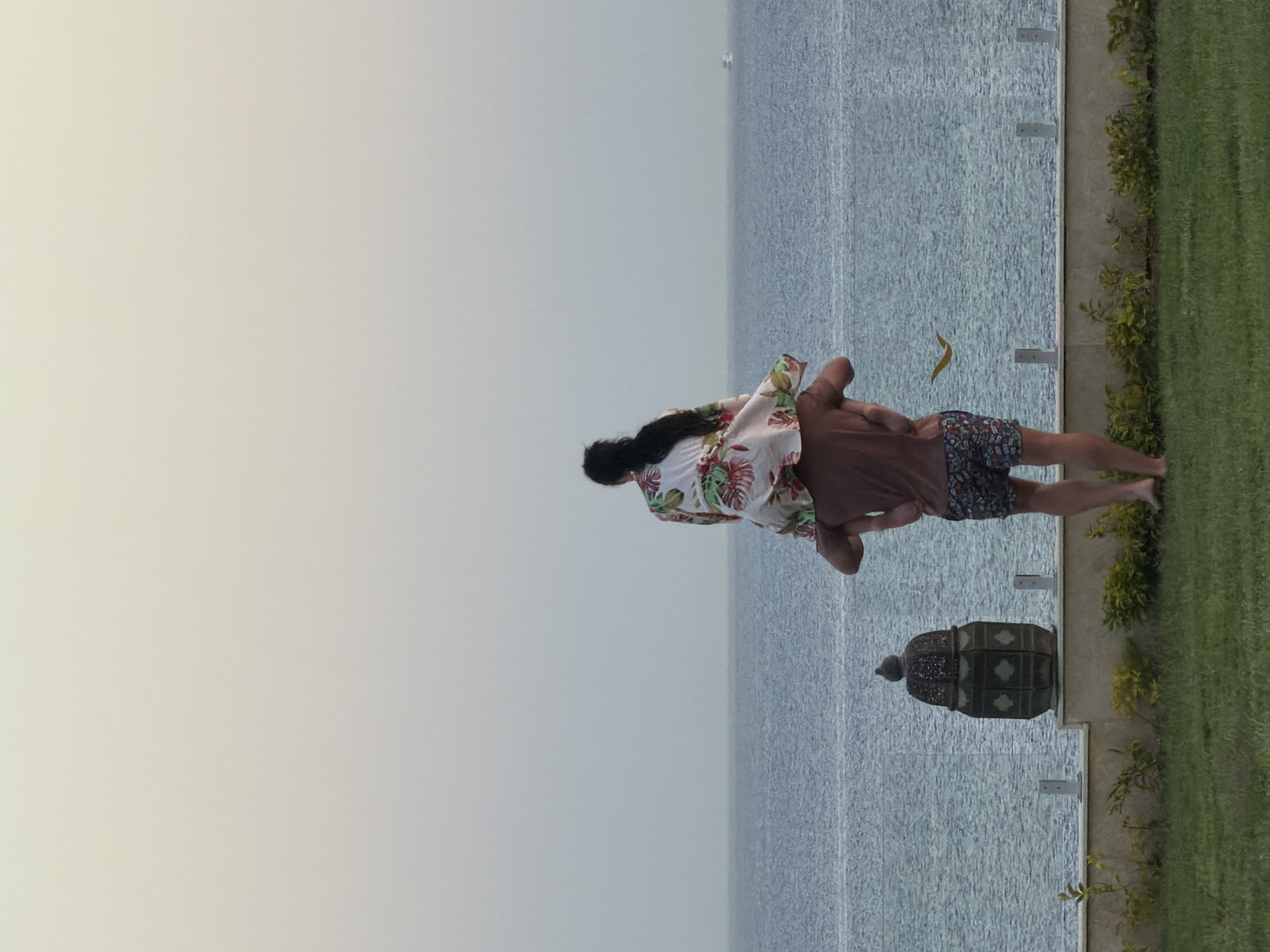 The image, if correctly oriented horizontally, captures two individuals, a man and a woman, standing by a calm body of water, likely the Red Sea, in a serene, resort-like setting in Sharm El Sheikh, Egypt. The scene appears to be set either in the early morning or late afternoon, characterized by soft, ambient light and an overcast or hazy sky.
The woman, with long dark hair, stands facing away from the viewer and towards the water. She is wearing a white, short-sleeved top adorned with a vibrant tropical floral pattern featuring red and green elements. The man stands closely behind her, partially obscured, wearing a plain brown short-sleeved top and dark, patterned shorts. Their posture suggests they are sharing a quiet moment, observing the expansive view.
They are positioned on a well-maintained grassy lawn, which transitions to a paved walkway or patio area. A transparent barrier, possibly a glass railing, separates this elevated area from the water's edge. In the foreground, on the paved section, a dark, intricately designed lantern or urn-shaped decorative object is visible. The water itself is a muted grey-blue, reflecting the soft light, and a single yellowish-brown leaf floats near the barrier. The distant horizon blends seamlessly with the light sky, indicating a peaceful, tranquil environment. No visible text is present in the image.