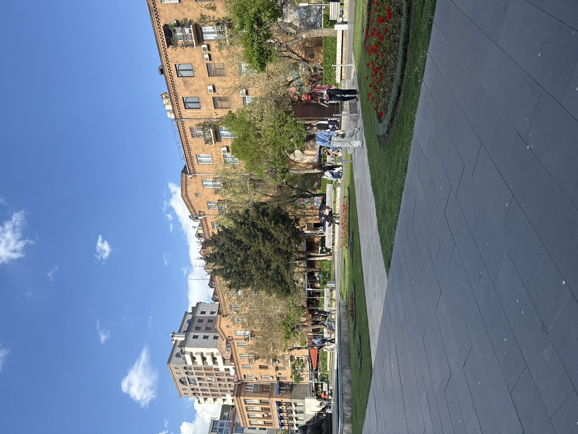 The image depicts a city scene in Yerevan, Armenia, likely along a pedestrian area or park adjacent to residential buildings. The sky is bright blue with scattered white clouds.
On the left side of the frame, several buildings are visible. These are primarily multi-story residential structures with a mix of architectural styles. The buildings are predominantly tan or brown. Some balconies and windows are discernible.
A strip of green space and a walkway occupies the central part of the image. People are seated on benches in the park. There is a flower bed with red flowers.
The right side of the frame is dominated by a dark gray paved surface.