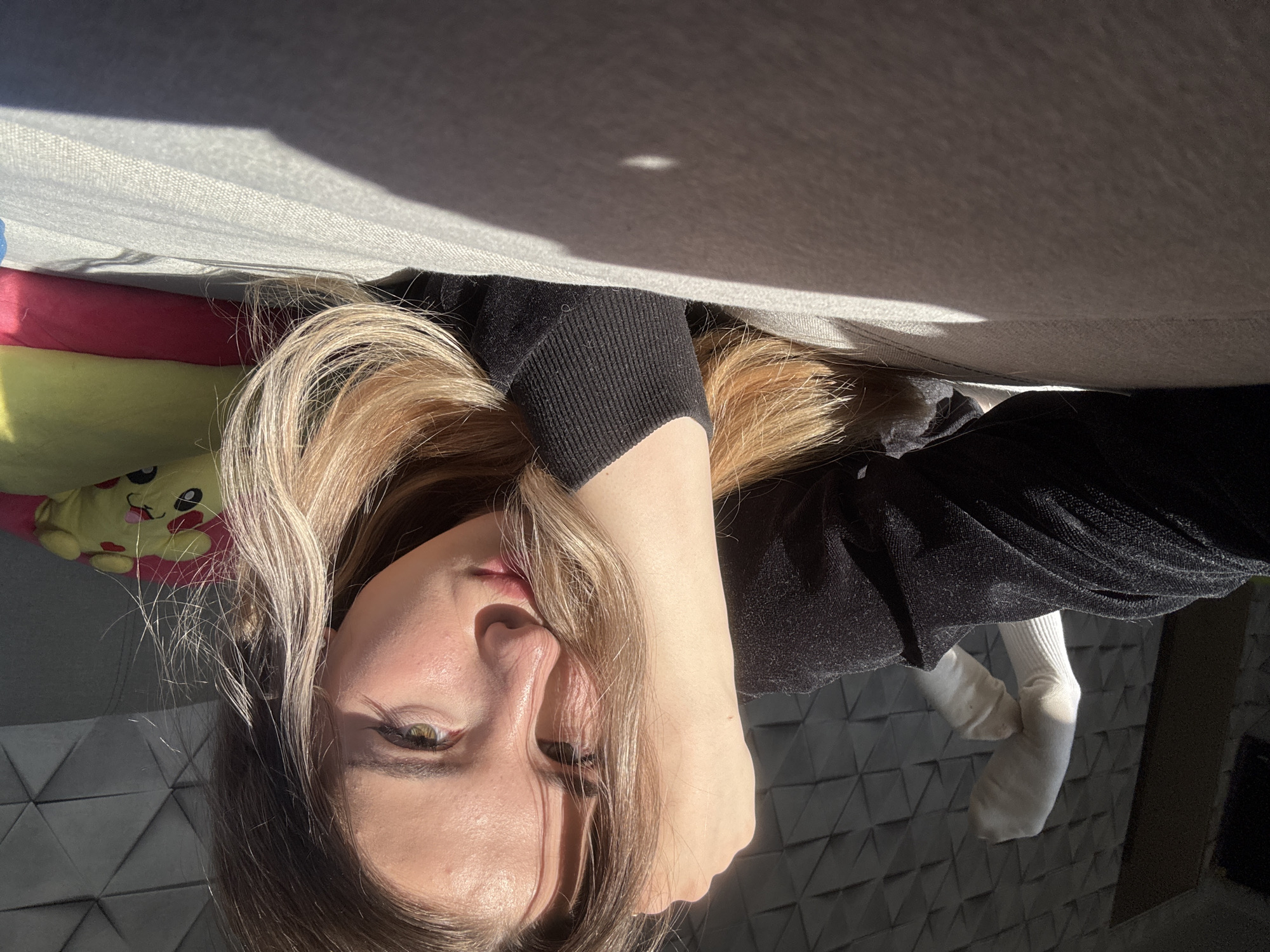 A young woman is depicted lying down, looking directly at the camera with a neutral expression. Her head is positioned on the left side of the frame, with her light brown hair, which has blonde highlights, partially covering her face. Her visible eye is green or hazel. She is wearing a black long-sleeved top with ribbed cuffs, and one arm is bent with her hand close to her face.
To her left, a bright yellow plush toy with a cartoon character face, resembling Pikachu, is partially visible, resting on a pink surface. Above the woman's head, a light-colored fabric surface, possibly a sofa or bed, stretches across the upper part of the image. Strong, bright sunlight illuminates sections of her hair and face, and creates distinct shadows on the fabric surface, indicating it is daytime. On the right side, a foot wearing a white sock is visible. The background features a dark wall adorned with a modern geometric pattern of repeating triangles. There is no visible text in the image. The scene suggests an indoor, relaxed setting, likely a home.