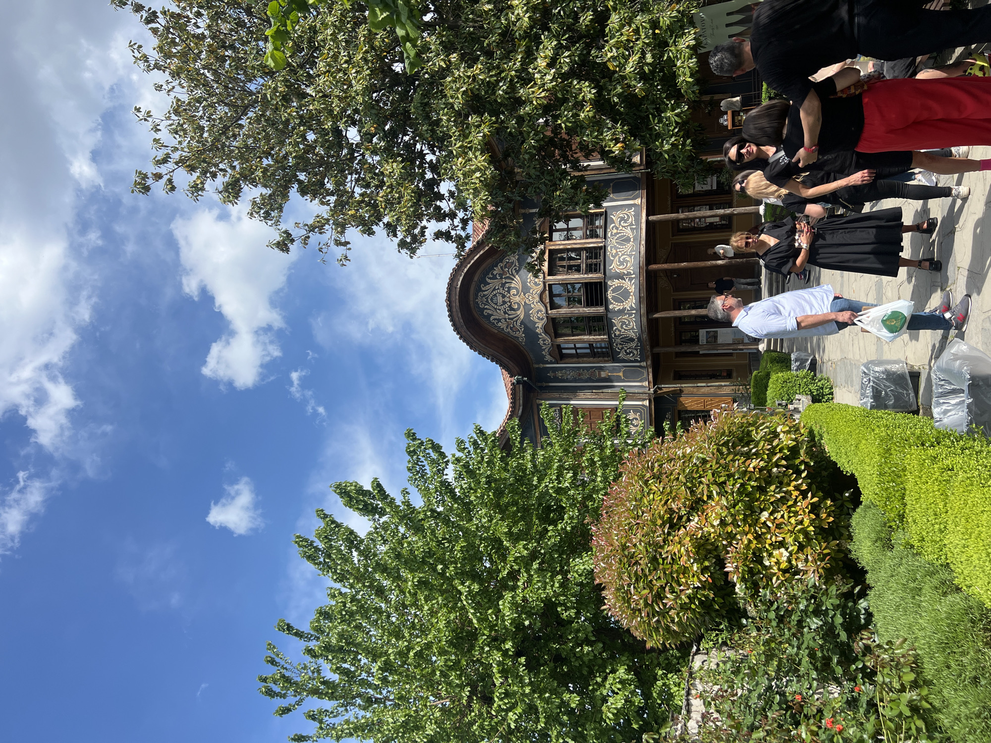 The image shows the facade of the Hindliyan House, a museum located in the Ancient Plovdiv area of Plovdiv, Bulgaria. The house is a two-story building with a distinctive facade featuring intricate wood carvings and floral patterns. The architecture is Balkan-Ottoman style. A group of tourists is standing outside, some of them wearing masks. A man in a white shirt and jeans is walking away from the house, holding a plastic bag. The sky is blue with scattered white clouds. Lush green trees and manicured bushes surround the house. The image appears to have been taken from ground level, looking up at the house.