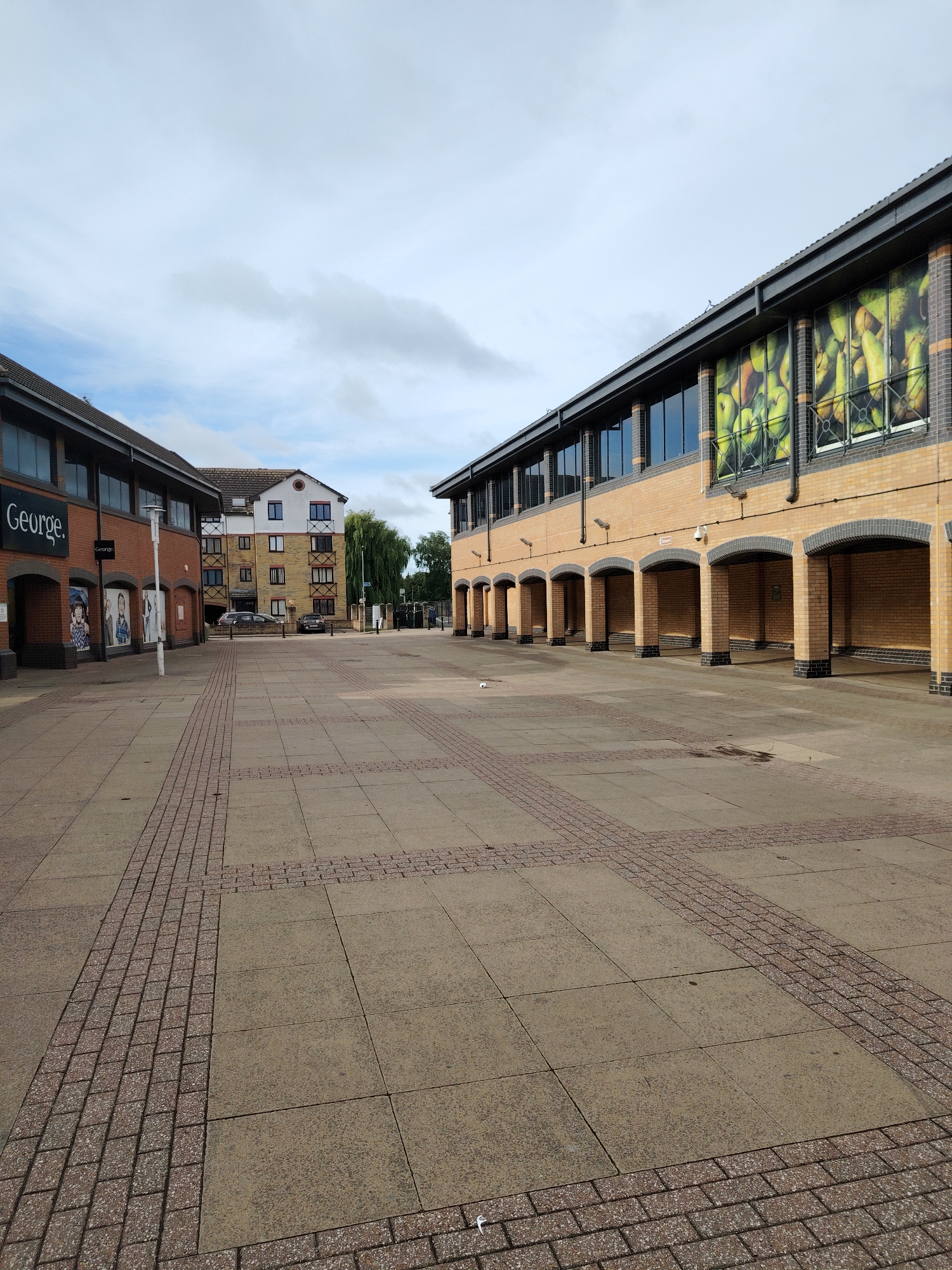This image captures a wide-angle view of a pedestrianized plaza or shopping precinct under an overcast sky. The scene is located within the Rivergate Shopping Centre, Viersen Pl, Peterborough PE1 1EL, United Kingdom.
On the left, a two-story commercial building with a light brown brick facade and a dark grey roofline extends into the foreground. This building features multiple arched entryways or display windows at ground level. A prominent white sign displaying the brand name "George." in a sans-serif font is mounted above an entrance.
On the right, a multi-story commercial building, also constructed with light brown brick and a dark grey roofline, mirrors the left structure's architectural style. Its ground level consists of a series of dark-arched recesses or covered walkways. The upper story features large windows, several of which are covered with a vibrant, large-scale graphic depicting a dense pile of green and yellow fruit, possibly pears or apples. Dark rain downspouts are visible on its facade.
The ground surface of the plaza is paved with large, light grey or tan concrete slabs. These slabs are interspersed with patterns of smaller, reddish-brown rectangular pavers that form distinct linear and cross-shaped designs, creating visual pathways across the open area.
In the background, situated behind the left commercial building and partially visible between the two main structures, stands a multi-story residential building. This building has a light beige brick base, a white upper facade, dark-framed windows, and a dark pitched roof. Several parked vehicles are visible in front of this residential structure. Further back and to the right, a large mature tree with dense green foliage is present.
A tall, cylindrical light-colored lamppost is positioned on the left side of the plaza, near the "George." building. Small pieces of white litter are scattered on the paving in the foreground and near the base of the right building. No individuals are present in the image. The overall lighting suggests an overcast daytime environment.