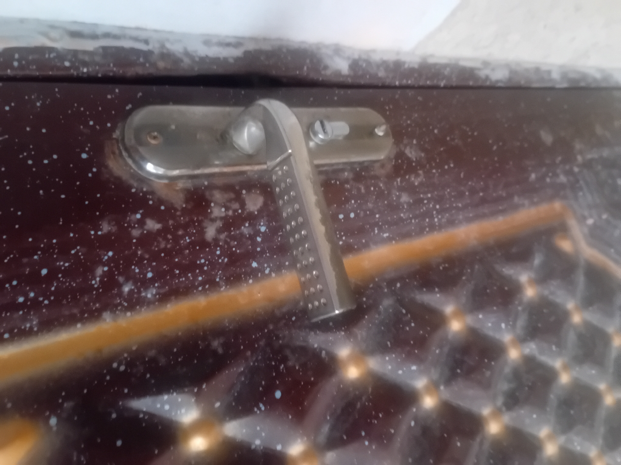 A close-up, eye-level shot shows the detail of a dark brown metal door with a metallic handle and lock mechanism. The door's surface is speckled with numerous small white and light blue dots, possibly paint splatter or debris. A vertical, golden-brown decorative element runs diagonally across the left side of the door, featuring a raised, diamond-shaped pattern with small golden spheres at the intersections. The door handle is made of brushed silver metal and has a textured grip. Below the handle is a keyhole with a silver metal surround. The right edge of the image shows a sliver of a white wall. The lighting is neutral and appears to be from overhead.