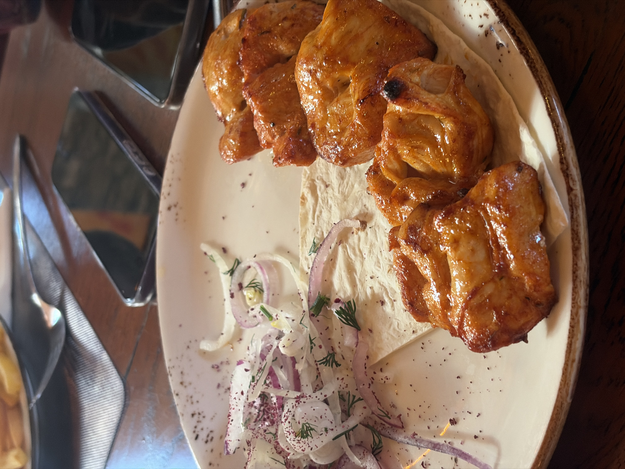 The image captures a close-up, top-down view of a plate of grilled food served on a wooden table, likely in a restaurant or cafe setting.
The main subject is a dish featuring several pieces of golden-brown, glossy grilled meat or poultry, possibly chicken shashlik or kebab, arranged on a bed of white flatbread (likely lavash). The meat has a slightly charred appearance in some areas, indicating grilling, and appears moist and tender.
Accompanying the meat is a fresh garnish composed of thinly sliced red and white onions, speckled with green herbs, which appear to be dill, and a reddish spice, possibly sumac, sprinkled over the onions and plate. The dish is served on a round, light-colored ceramic plate with a distinctive speckled brown rim, suggesting a rustic or artisanal design.
In the blurred background and reflections on the left side of the image, parts of other table settings are visible, including the metallic gleam of silverware (forks and knives) and what appears to be the edge of another plate or bowl. There's also a reflection of a dark, rectangular object with indistinct white lettering, but no specific words are clearly readable. The table surface is made of dark wood, contributing to a warm, inviting atmosphere. The lighting suggests an indoor setting, possibly evening or artificial light. No people are visible, and no specific location cues or emotions are discernible from the image itself, beyond the provided context of Saint Petersburg, Russia.