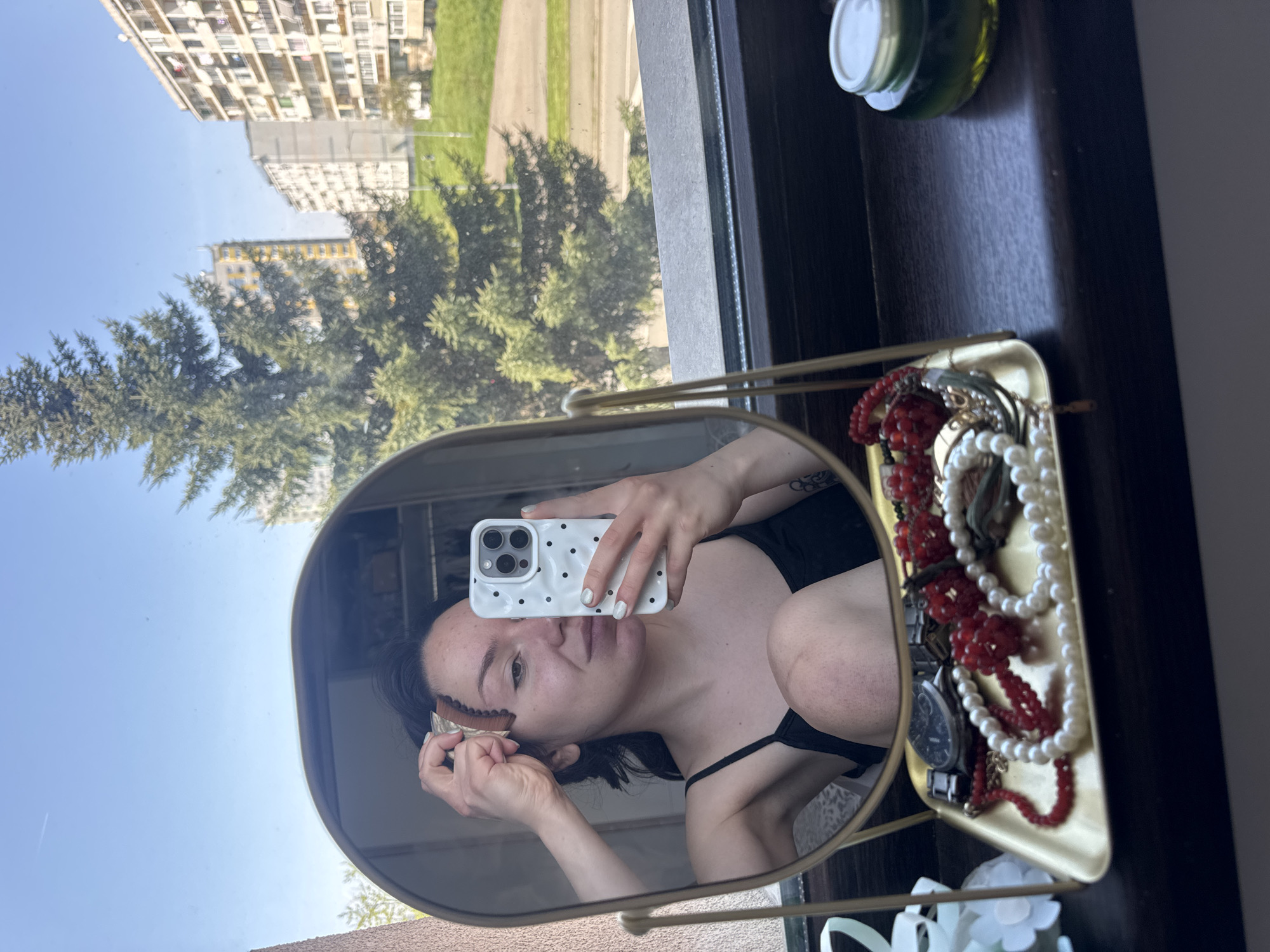 The image is a selfie taken in a mirror. The subject, a woman with dark hair, is holding a cell phone with a white case. She is applying makeup to her face with a brush. A gold tray with jewelry, including red and white beaded necklaces and a watch, is visible next to the mirror. In the background, seen through a window, is an exterior view of buildings and trees. The buildings appear to be residential apartments. Based on the architecture and landscape visible, the scene is likely located in Burgas, Bulgaria.