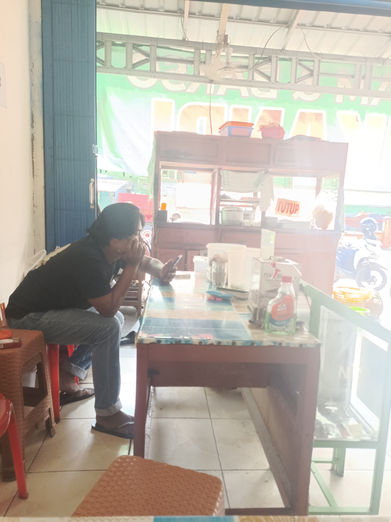 A man with dark hair, wearing a black t-shirt and blue jeans, is sitting on a brown plastic chair in an open-air establishment in Sangatta, Indonesia. He is looking at his mobile phone, held in his right hand, with his left hand supporting his chin. He is positioned at a table with a colorful patterned surface, which also holds several items including a bottle of cleaning fluid, a box, and various containers. Behind the man and the table, a food stall structure is visible, with a green banner displaying large white lettering. A motorcycle is parked outside the stall. The environment appears to be a casual eatery or street-side vendor.