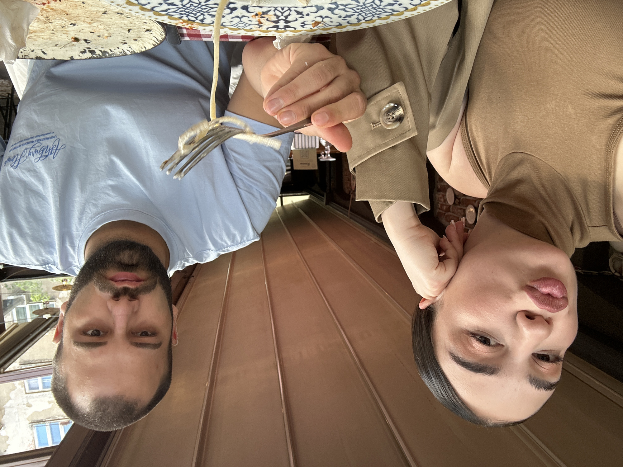 Two individuals, a male and a female, are positioned looking downwards towards the camera. The setting appears to be a restaurant or dining establishment, as indicated by a table with food and dining ware visible at the top of the frame. A fork is present, holding what appears to be pasta. The male individual has a beard and wears a light blue t-shirt. The female is wearing a tan or brown colored garment. The background features a hallway with wood paneling. Based on the window exterior, it is in the city of Sofia, Bulgaria.