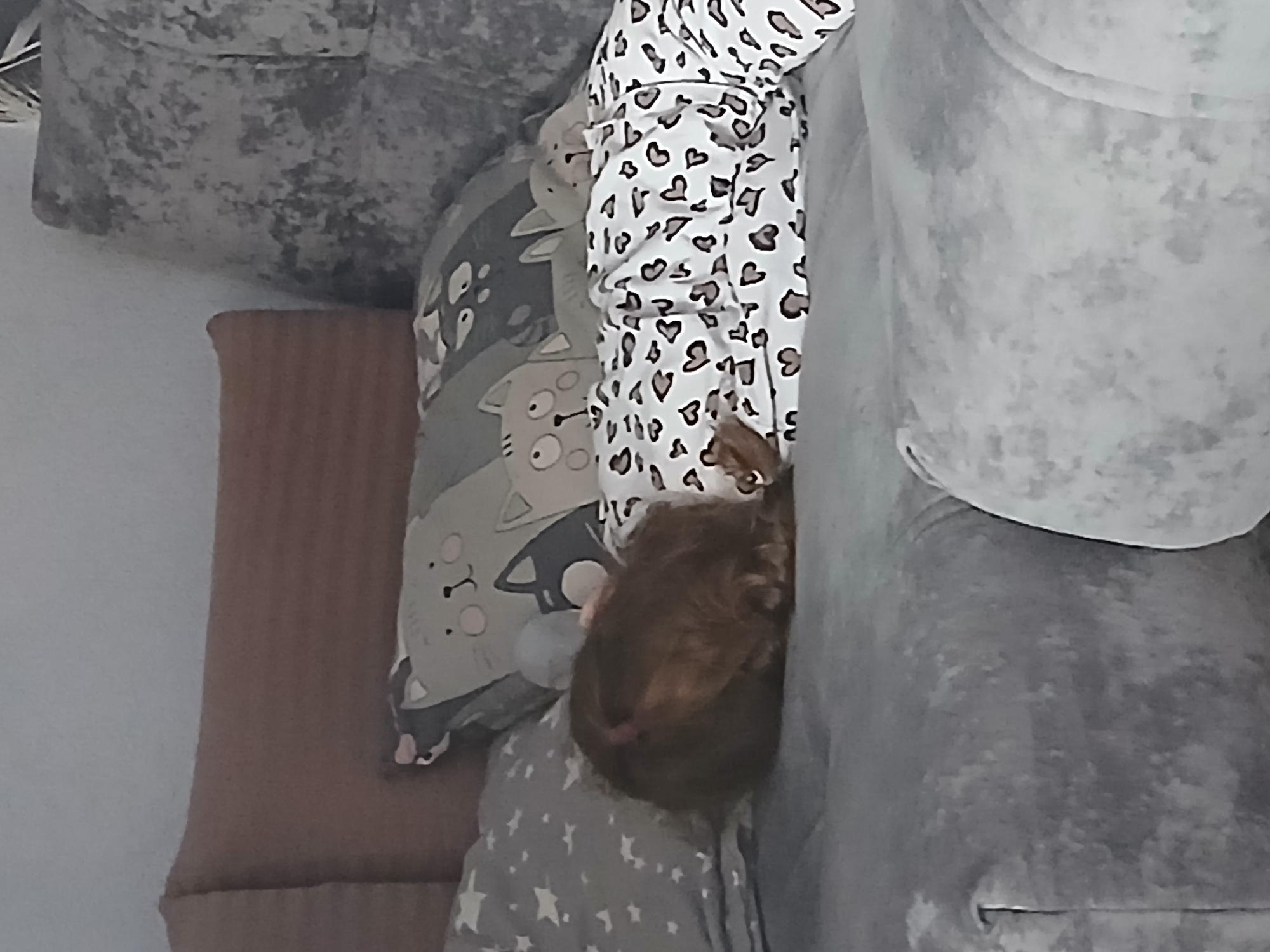 A child lies face down on a sofa, wearing white pajamas with brown heart shapes. The child's brown hair is visible. The child rests on a gray pillow with cat cartoons and a gray pillow with white stars. The sofa is gray. A brown cushion is adjacent to the pillows. The location is Марковский, Russia.