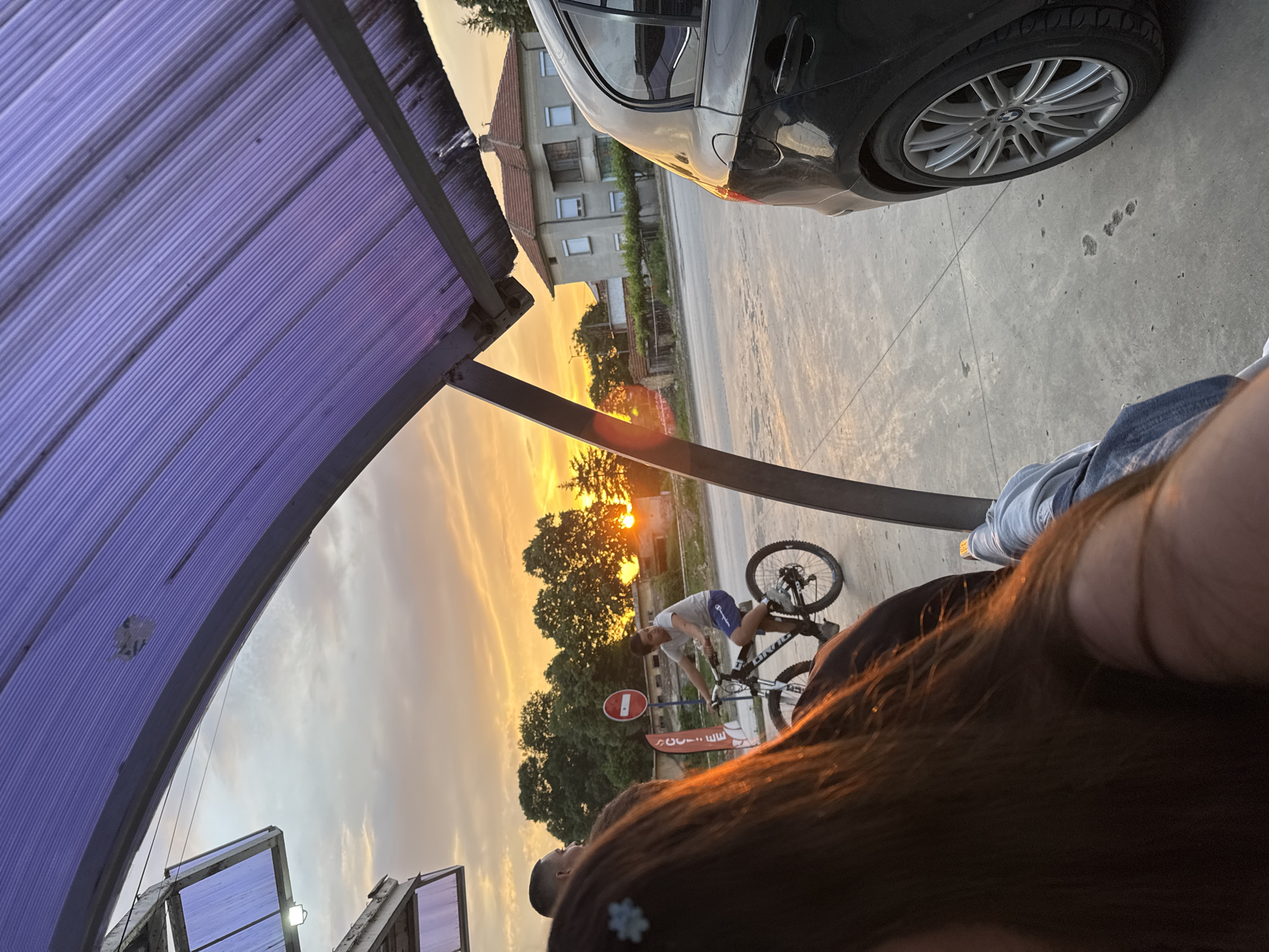 The image depicts a sunset scene in Dolna Oryahovitsa, Bulgaria. A male is riding a mountain bike. A stop sign is visible in the background. The photo is taken under a purple corrugated metal roof. A portion of a dark-colored car is visible in the upper right corner. Another person is visible in the foreground, with long brown hair partially obscuring the view. The sky is a mix of orange and yellow hues.