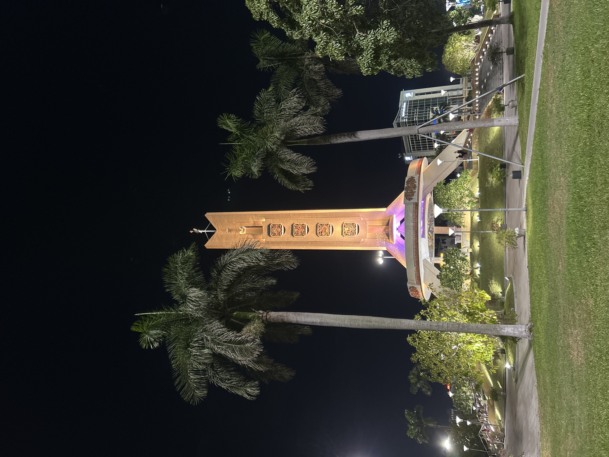 This is a night photograph of the Dragon Bridge in Da Nang, Vietnam. The bridge is illuminated with colorful lights, and palm trees are visible in the foreground. The sky is dark, indicating that it is nighttime. The bridge appears to be a popular tourist destination, as there are several people visible walking on the bridge and in the surrounding area.