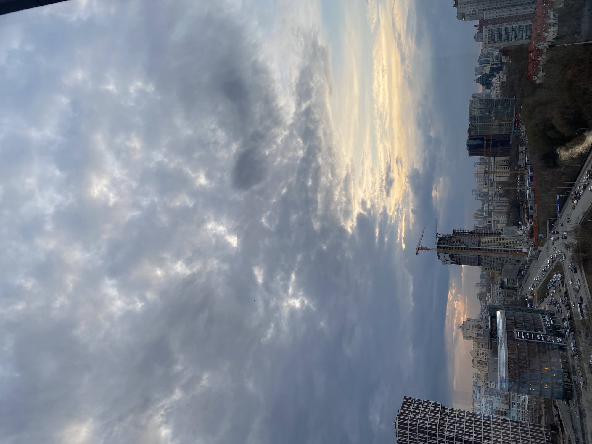 The image captures a cityscape under a cloudy sky. Buildings of varying heights and architectural styles form the urban landscape. Construction cranes are visible near some buildings, indicating ongoing development. A busy road with vehicles runs alongside the buildings. The photo was taken in Yekaterinburg, Russia.