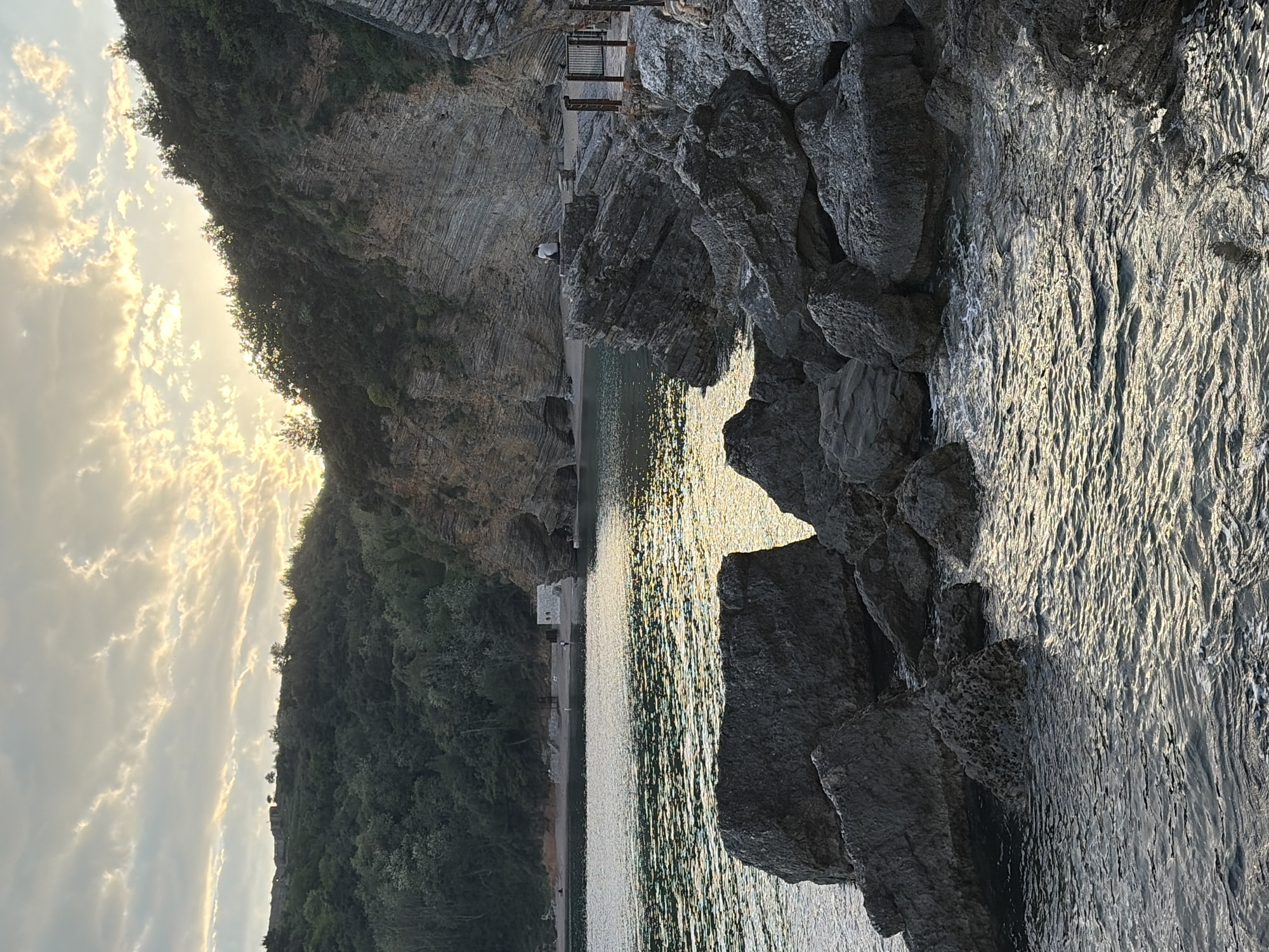 The image shows a coastal view in Budva, Montenegro, captured during daytime under a partly cloudy sky with sunlight reflecting on the water. Jagged rocks are prominently featured on the right side of the frame, while a cliff face covered with sparse vegetation is visible on the left. In the distance, a narrow beach stretches along the base of the cliff, with some structures and a pedestrian bridge visible. A single person is present on the beach. The water appears calm, with a textured surface created by the sunlight.