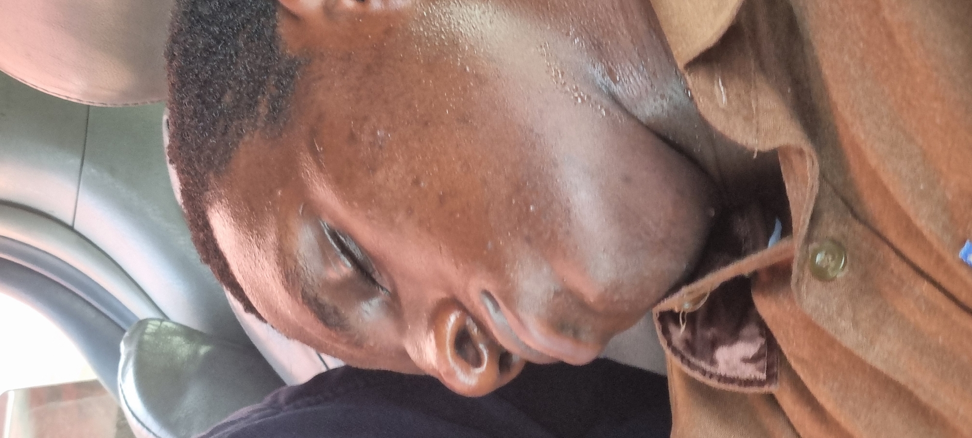 A medium shot depicts a Black male resting in what appears to be the front passenger seat of a vehicle. His head leans against the seat, close to the interior roof lining. The man has short, cropped hair, and his skin appears somewhat shiny with moisture. He is wearing a brown shirt or jacket with a button visible. The background shows parts of the vehicle's interior, including the car door and headrest. The location, Satellite Town, Nigeria, is based on contextual information not visible in the image itself.