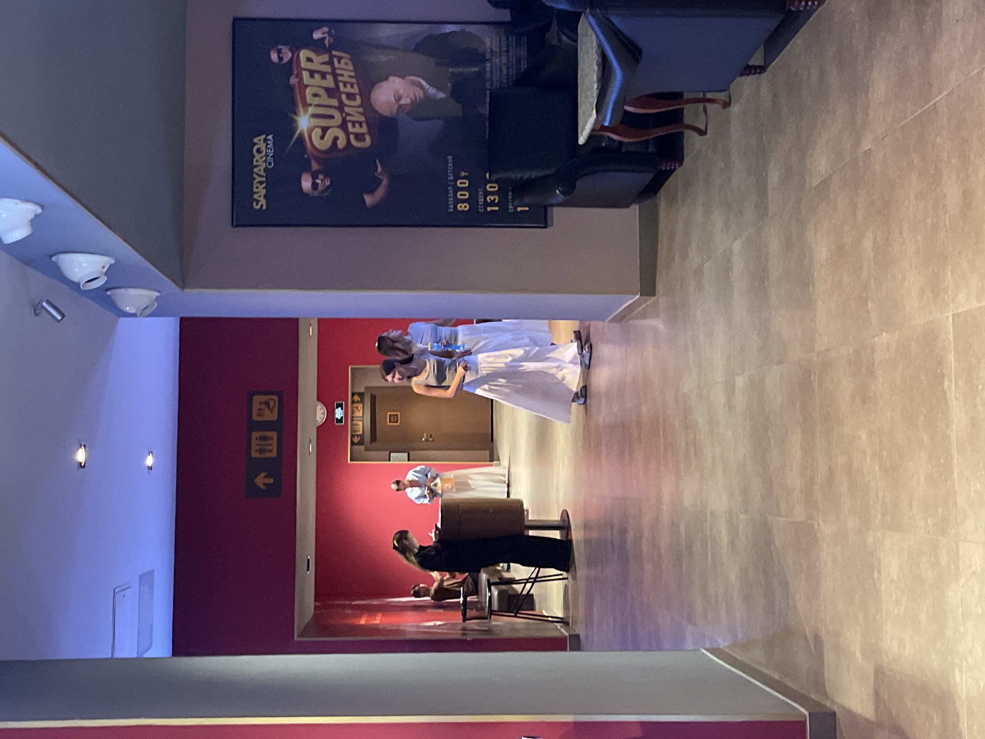 The image depicts an indoor scene, likely a cinema lobby, with a focus on a hallway leading to various rooms. The walls are painted in a vibrant pink, and the floor is tiled in a neutral beige.
In the background, a person in a black dress is standing at a reception desk, possibly attending to a customer. To the right, two women in white skirts and tops are conversing, while another woman in a grey tank top and white skirt walks past. Several doors are visible along the corridor, some with signage indicating amenities like restrooms. A movie poster for "SUPER СЕЙСЕНБ!" is prominently displayed on the right wall, featuring images of actors and pricing information for tickets.
In the foreground, plush leather sofas and an armchair suggest a waiting area. The lighting is a mix of ambient ceiling lights and spotlights, casting a warm glow on the scene. The overall impression is one of a functional and somewhat casual entertainment venue. The "SARYARQA CINEMA" text on the poster and the Cyrillic script suggest the location is likely in Kazakhstan, and more specifically, Almaty.