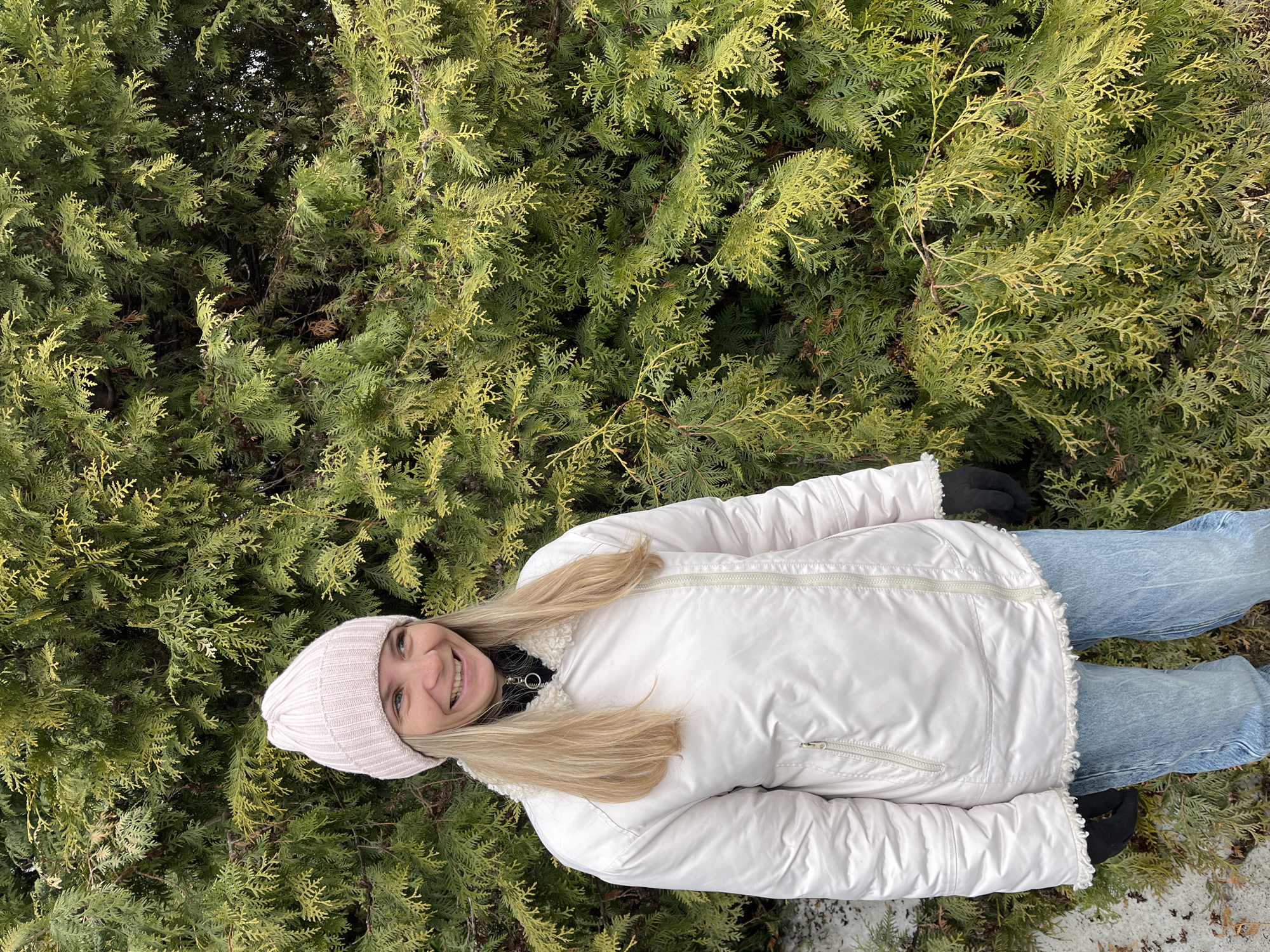 A woman wearing a light pink knit hat, a white winter coat with a furry trim, blue jeans, and black gloves is lying on the ground next to a dense evergreen shrub. The woman is smiling and looking at the camera. Based on available information, the location is the city of Chaikovsky, Russia. The ground is partially covered with patches of snow. The evergreen shrub fills much of the background.