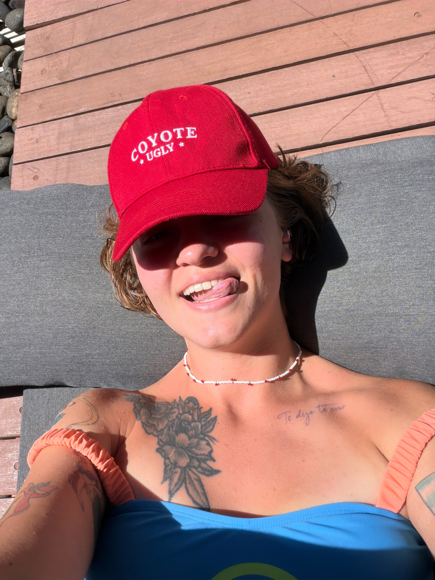 A woman wearing a red baseball cap that says "COYOTE UGLY" and a blue bikini top is lying on a gray lounge chair. She is sticking her tongue out and looking at the camera. She has several tattoos on her arms and chest. The scene is outdoors, with a wooden deck in the background and sunlight casting a shadow.