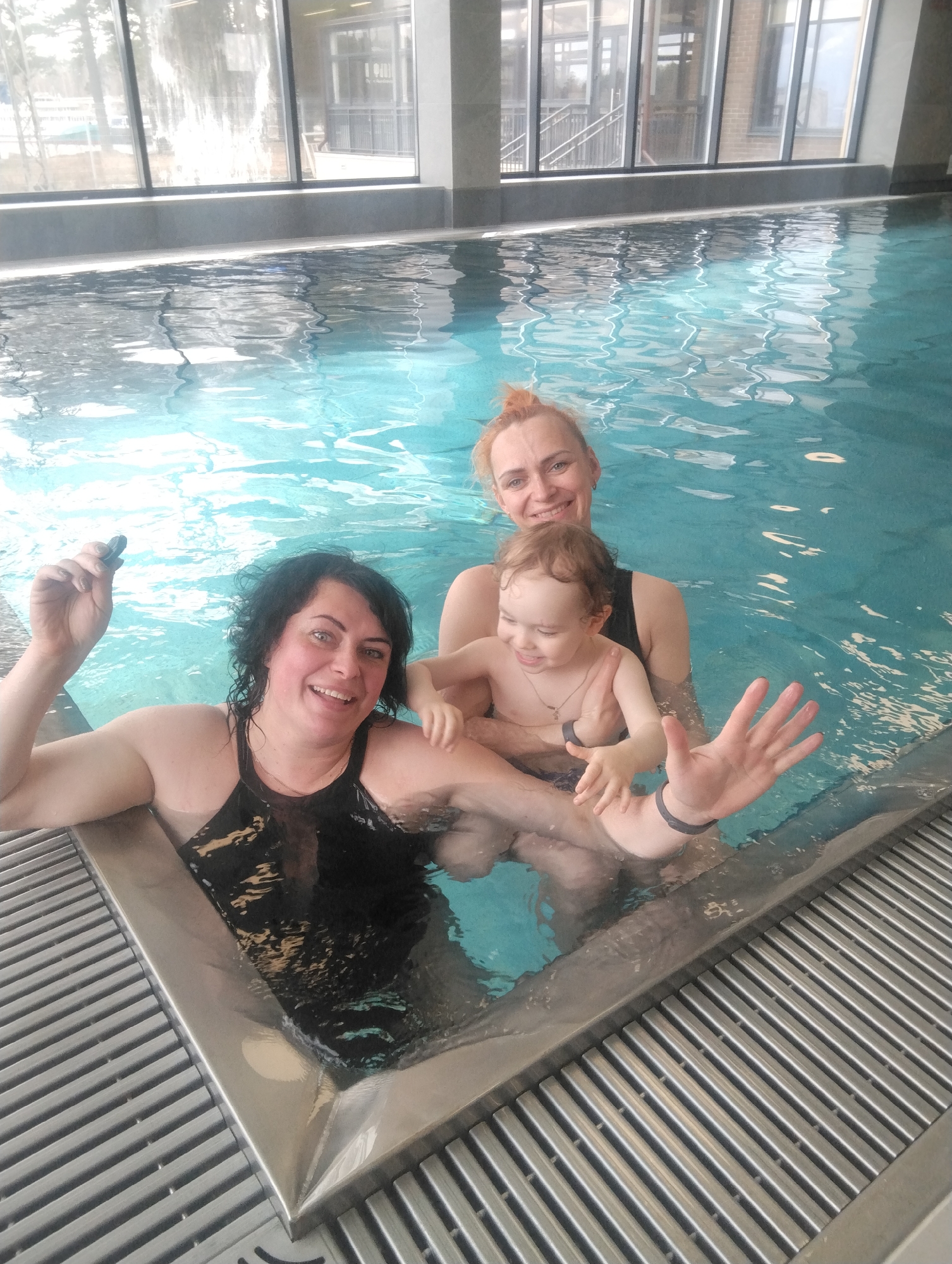 An indoor swimming pool scene captures three individuals, two women and a young child, joyfully immersed in clear blue water. All three are smiling brightly at the camera, radiating happiness and amusement.
The woman on the left has dark, curly hair and wears a black swimsuit. She raises her left hand, holding a small, dark, smooth object, and extends her right hand with fingers spread in a lively gesture. A dark band adorns her right wrist. The woman on the right, with reddish-blonde hair pulled back and wearing a dark swimsuit, supports the young child between them. The child, with light brown hair, is laughing, wearing dark swim bottoms and a necklace.
Behind the pool, large, multi-paned windows reveal reflections of trees and what appears to be distant buildings, indicating it is daytime. The pool itself features a modern design with a metallic edge and a surrounding grey grate system. The water is gently rippled. The setting suggests a contemporary public pool, spa, or hotel facility in Chaikovsky, Russia. There is no visible text in the image.