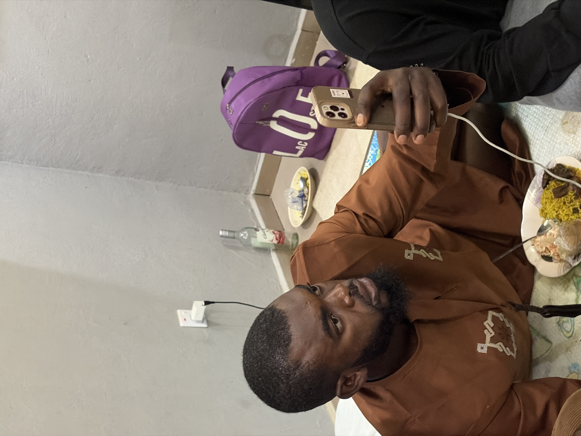 A man with a beard is holding a smartphone connected to a charging cable. He is wearing a brown robe with white geometric embroidery. To his right is a plate of food. Next to him is a purple bag with white lettering. Behind the bag is a small bowl, and further back is a glass bottle. On the wall to the left, there is an electrical outlet.
Given the context and details like the style of clothing, a possible location for this scene could be a residential setting in Jos, Nigeria. The specific location cannot be determined from the image alone.