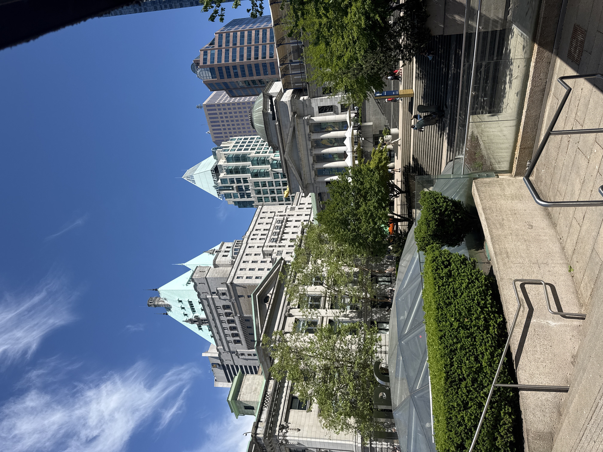 The image depicts the area around the Vancouver Art Gallery in downtown Vancouver, Canada. Several buildings of varying heights and architectural styles are visible against a bright blue sky with some wispy clouds. The buildings include a classical-style building with columns and a dome and modern skyscrapers. In the foreground, there are stairs, landscaping with bushes, and metal railings. People are walking on the steps. The image appears to have been taken from a slightly low angle, tilting upwards.