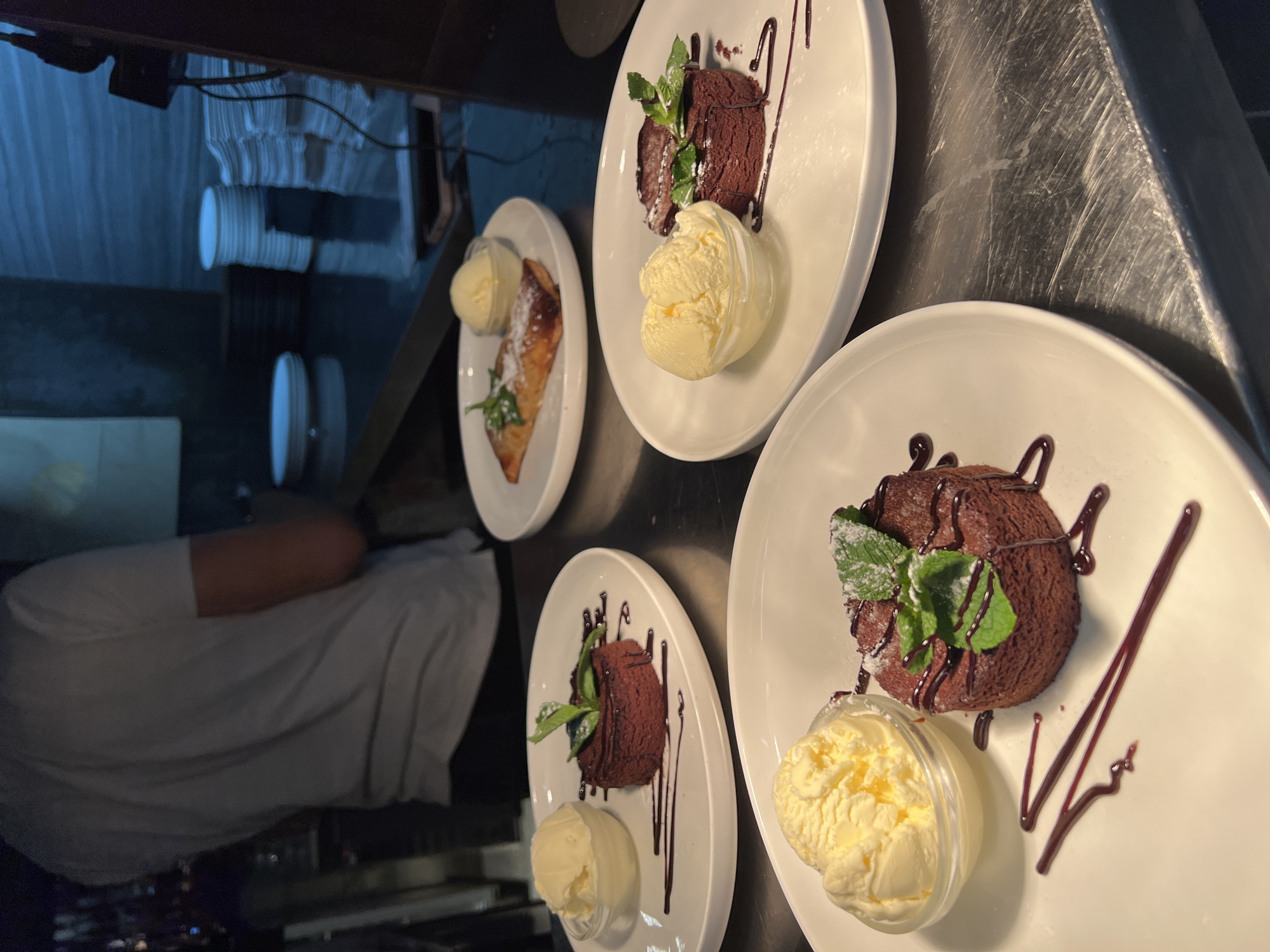 The image shows a culinary setup, likely within a restaurant or kitchen environment. There are three plates of chocolate desserts, each garnished with mint and chocolate sauce, accompanied by a scoop of ice cream served in a small transparent container. One plate displays a pastry, dusted with powdered sugar, also with a scoop of ice cream. There is a person wearing a white shirt standing next to the plates. In the background, there are stacked plates and lighting fixtures. The reflective surface of the countertop suggests it could be stainless steel. The absence of contextual details prevents pinpointing the location, or any particular city. No information is provided to indicate the setting being in Budva, Montenegro, or any city at all.