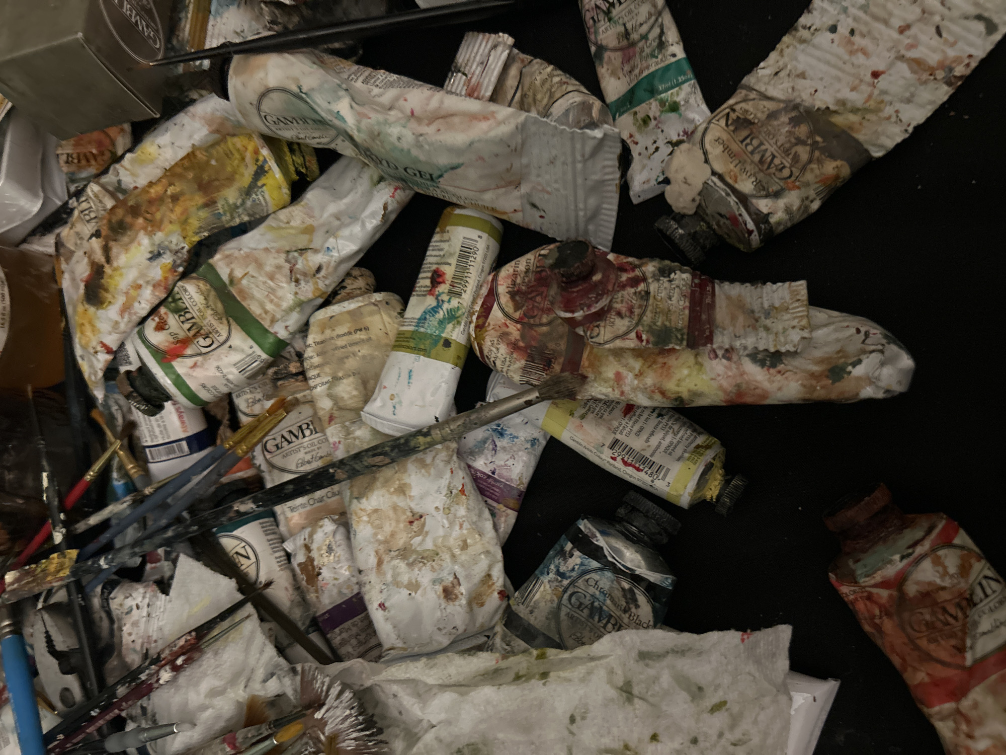 This image shows a collection of used art supplies, primarily oil paints and paintbrushes. The tubes of paint are mostly from the Gamblin brand and show signs of wear and use, with paint splatters and squeezed tubes. The color names visible include Sap Green, Teinte Chair Cau, Chroma Black, Raw Umber, and Alizarin Crimson. There are a variety of paintbrushes scattered amongst the paint tubes. Some appear clean while others have dried paint on the bristles. There's also a box of Gamblin paint. The image doesn't provide any information about location but I am told to answer "Cape Coral, United States of America".