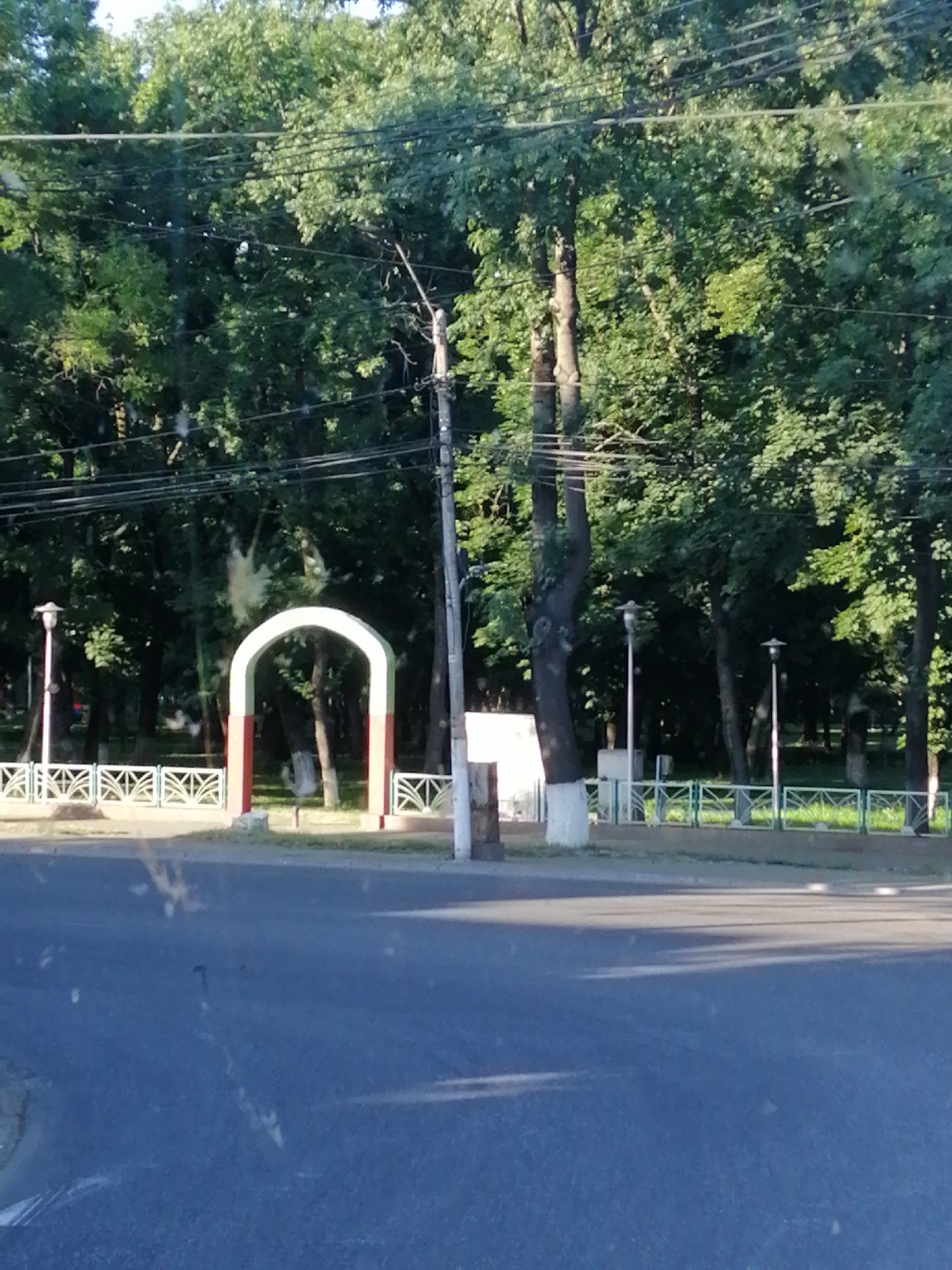 This media file depicts an outdoor scene featuring a park entrance. The central object is a white arched gateway with red and green accents, marking an entrance into a tree-lined area. In front of the gateway, a decorative white metal fence runs horizontally across the frame, interspersed with evenly spaced posts. Beyond the fence, various trees with lush green foliage dominate the background, creating a sense of depth and natural beauty.
Several lampposts are visible, two of which are positioned near the fence and gateway, suggesting a public park or garden. A utility pole with multiple wires is also prominent, cutting vertically through the scene, indicating the presence of infrastructure. The ground in the foreground appears to be asphalt, suggesting the perspective is from a street or pathway bordering the park.
The time of day appears to be late afternoon or early evening, as indicated by the shadows cast across the asphalt and the warm lighting filtering through the trees. The weather seems pleasant, with clear skies and abundant greenery, implying a tranquil and possibly warm day. No people are visible in the frame, lending a sense of quietude to the scene. The overall atmosphere is peaceful and inviting, highlighting the natural beauty of the park. No text is visible in the image.