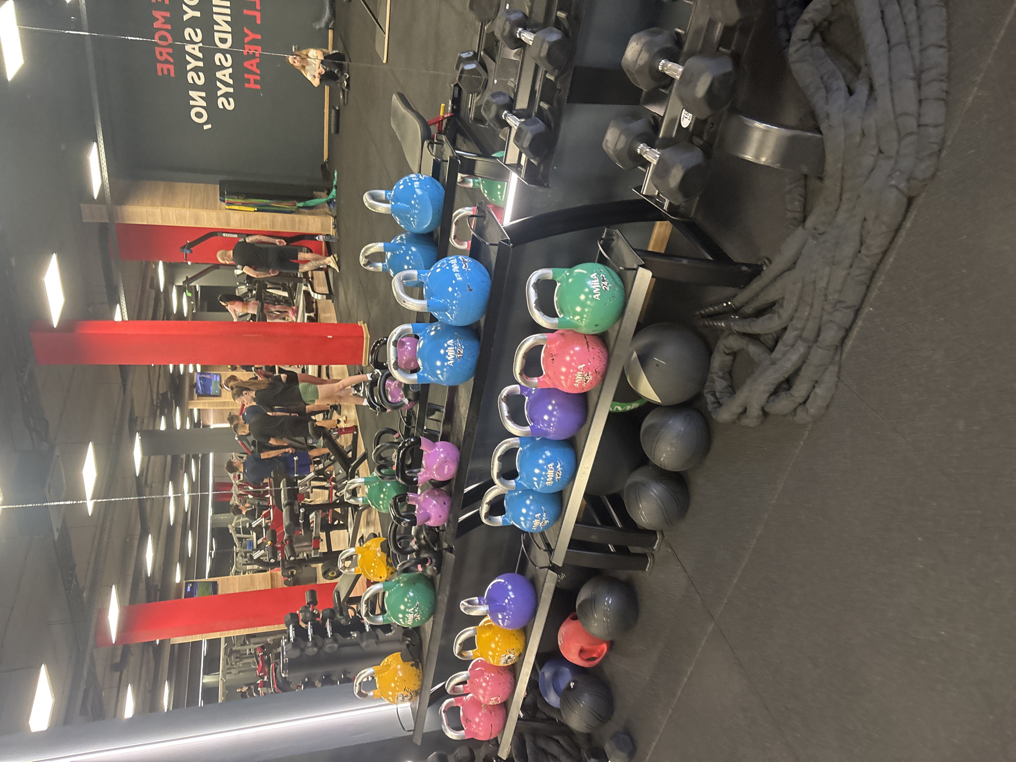 The image captures the interior of a modern gym or fitness center, filled with various exercise equipment and several people. The scene is dominated by racks of colorful kettlebells and black dumbbells, arranged neatly on multiple shelves. The kettlebells display a range of weights and colors, including blue, green, pink, purple, and yellow, with the brand name "AMILA" visible on some. Alongside these, black medicine balls or slam balls are also stacked. A large coiled black battle rope rests on the floor next to a dumbbell rack.
The gym features a dark floor and bright, rectangular overhead lights, which are reflected extensively in large mirrors lining the walls. These reflections show more of the gym's interior, including various exercise machines and additional individuals working out. In the reflected view, a person with blonde hair is seen sitting on the floor, while a man with tattoos on his leg is actively using a piece of gym equipment. Other indistinct figures are visible further in the reflections, suggesting a busy atmosphere.
On a dark wall, a motivational text is visible, partially directly and partially reflected. When properly oriented and deciphered, it reads: "NO MORE EXCUSES. YOU ARE YOUR ONLY LIMIT. YEAH!" The lighting suggests it's daytime or early evening indoors. The context provided indicates the location is Plovdiv, Bulgaria.