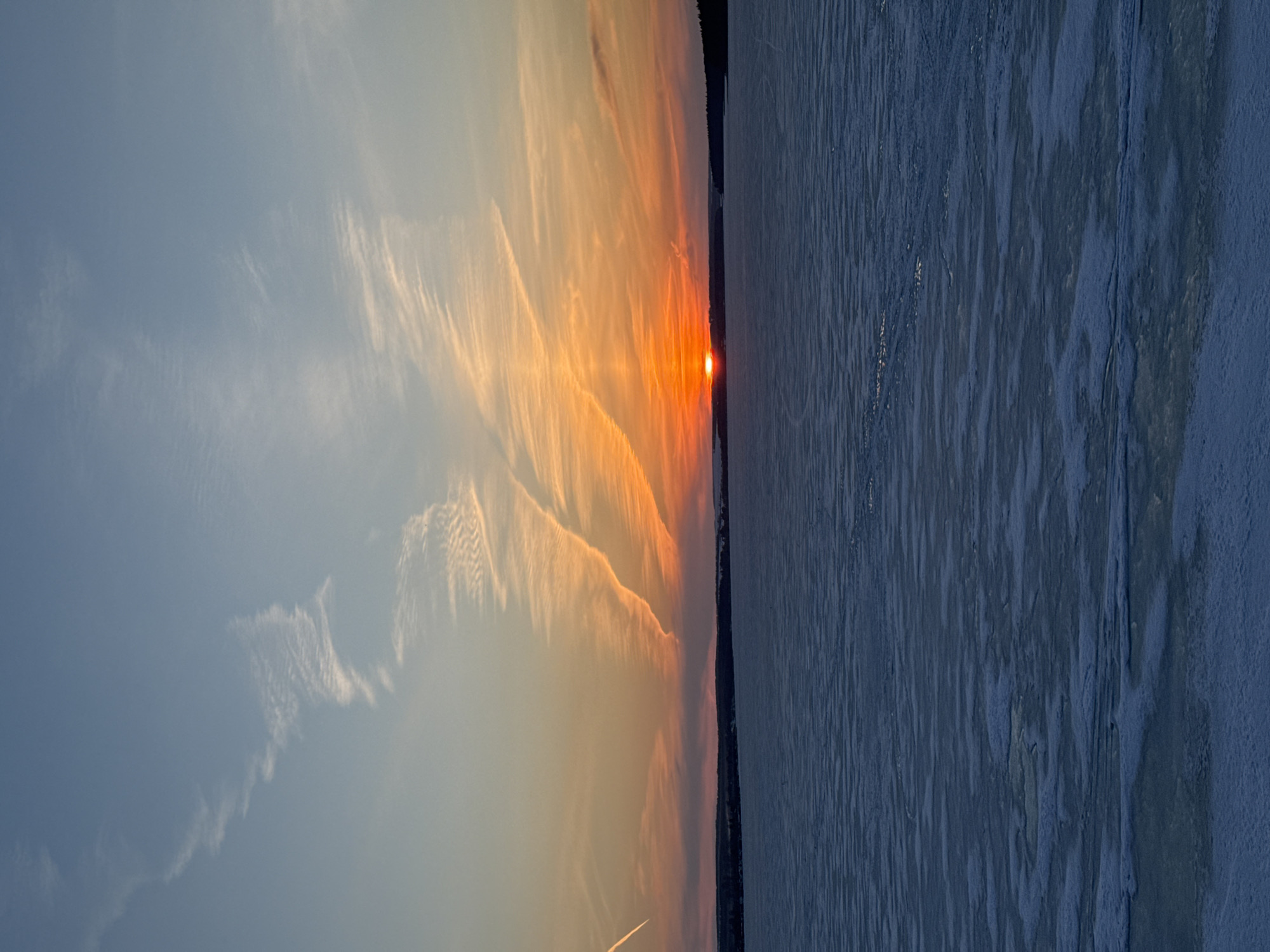 The image depicts a sunset over a body of water, likely a frozen lake or river. The sky is divided into sections of blue and orange hues, with clouds and contrails present. The sun is visible as a small, bright point on the horizon. The water's surface is blue and partially covered with what appears to be ice.
The image's details suggest the location may be in Votkinsk, Russia, due to the landscape and the presence of frozen bodies of water common in the region during certain times of the year. However, there are no identifying landmarks or features that definitively confirm the location.