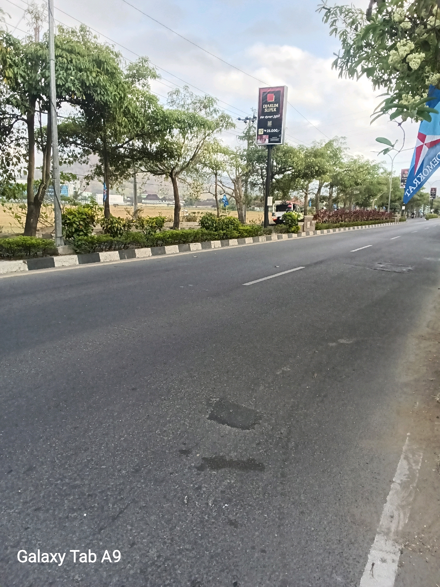 The image shows a street in Trenggalek, Indonesia. The street is paved with asphalt and has a white line marking the lane. On the left side of the street, there are trees and bushes. On the right side, there is a sidewalk and more trees. In the background, there are buildings and a sign that says "DIARUM SUPER". The sky is cloudy. There is a white van parked on the left side of the street, and a blue flag with white stars is visible on the right. The bottom left corner of the image has a watermark that reads "Galaxy Tab A9".