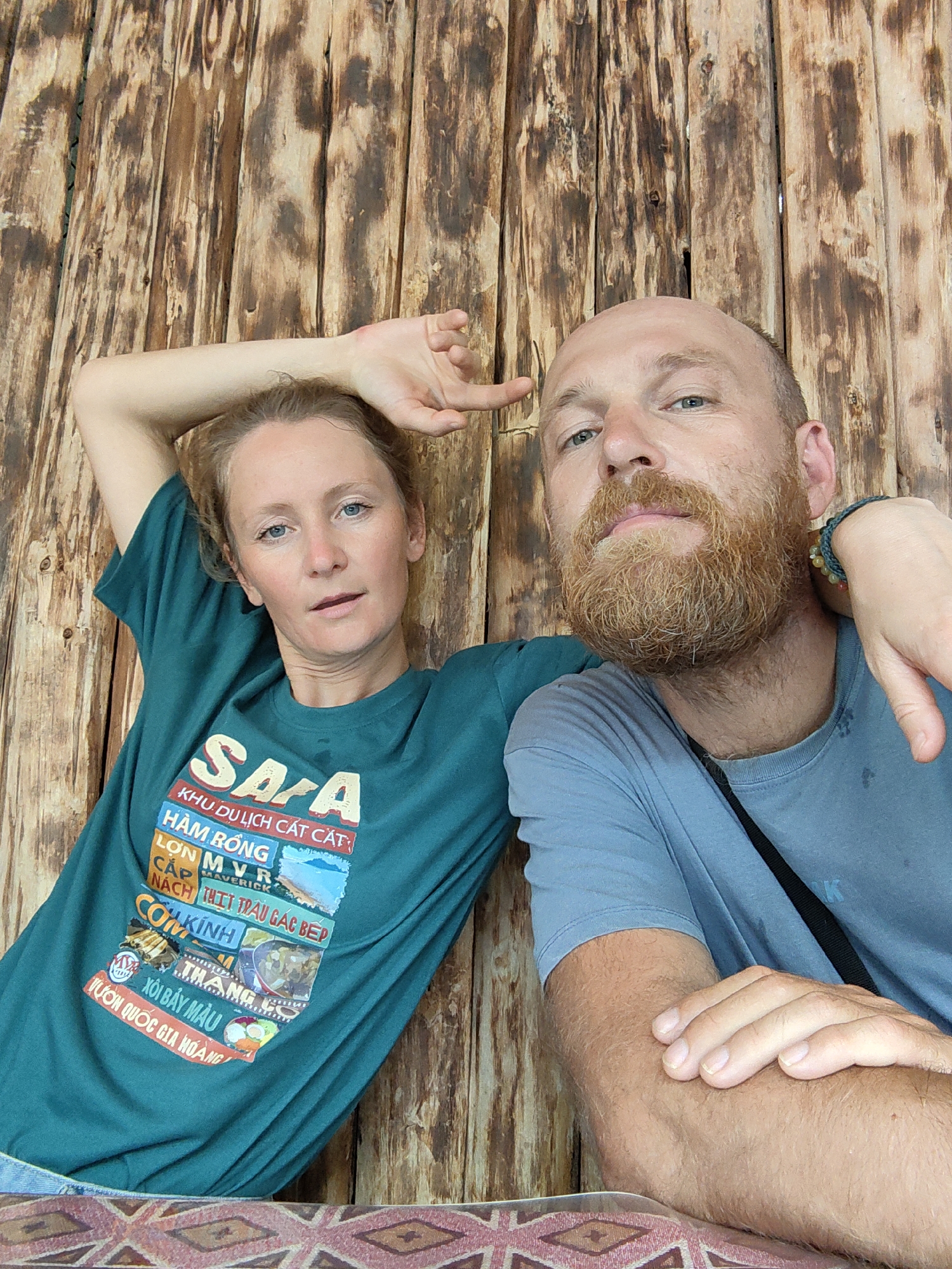 A medium, eye-level shot depicts two adults seated indoors, positioned against a rustic wooden wall.
The woman, fair-skinned with blonde hair, is wearing a teal t-shirt with multiple colorful images. She has one arm raised, hand near her head. Her expression is neutral.
The man, fair-skinned and bald with a reddish beard, is wearing a light blue t-shirt. He is slightly in front of the woman. He has one arm resting on the table in front of him and the woman's hand on his shoulder. His expression is a small frown.
The wooden wall consists of vertical planks.
The bottom of the frame shows a patterned tablecloth, likely covering a table.
The t-shirt has the following text: Sapa KHU DU LỊCH CÁT CẮT HÀM RỒNG LỢN MVR CẮP MAVERICK NÁCH THỊT TRÂU GÁC BẾP COKÍNH THANG XÔI BẢY MÀU VƯỜN QUỐC GIA HOÀNG.
Given the text "Sapa," it is inferred the photograph was taken in the city of Thành phố Hà Giang, Vietnam.