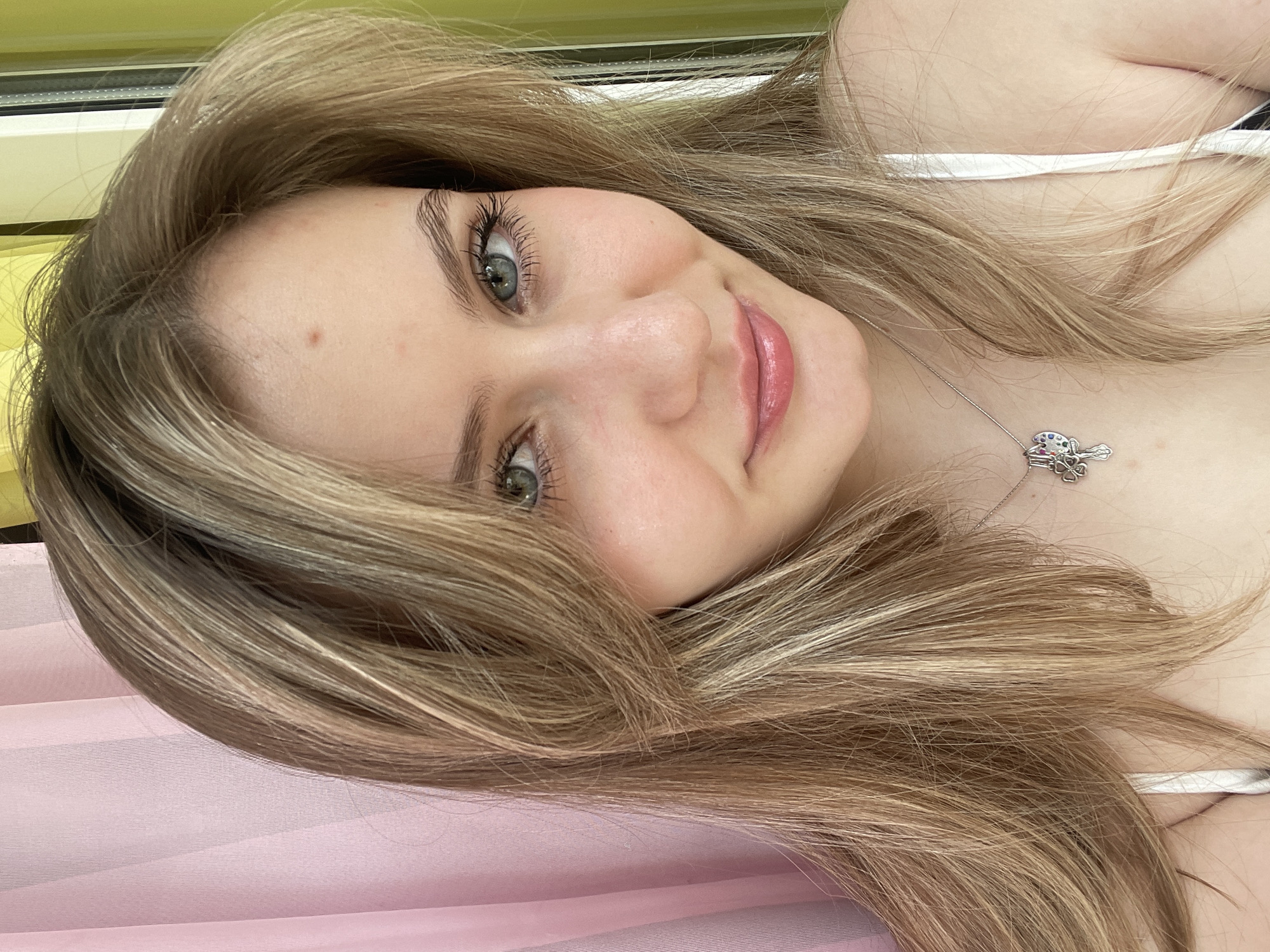 A young woman with blonde hair and blue eyes is looking at the camera. She is wearing a white tank top and a silver necklace with a clover charm. The background is a pink curtain and a green curtain.