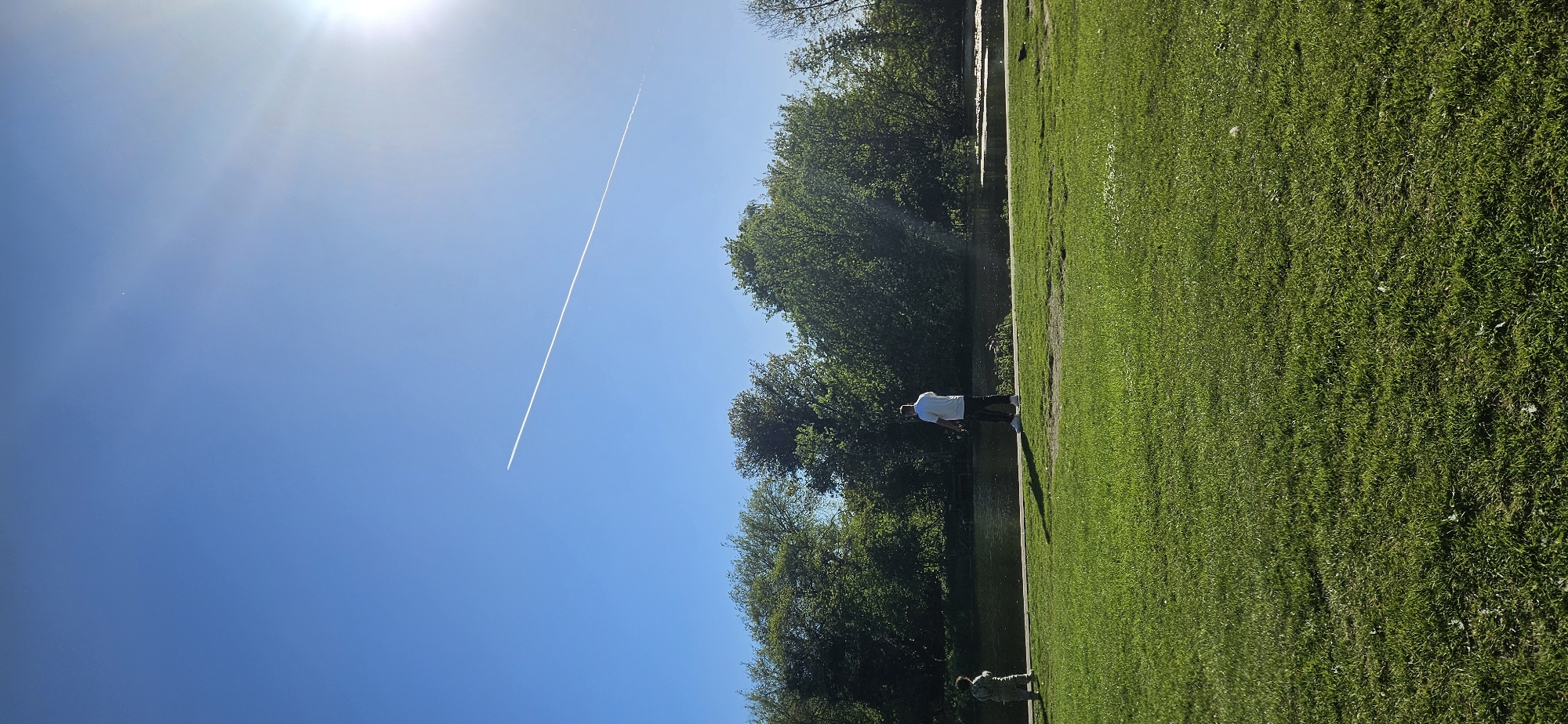 This image, correctly oriented, depicts a serene outdoor scene, likely a park or canal bank, bathed in bright midday sun. The upper portion of the frame is dominated by a clear, deep blue sky. The sun is prominently visible in the upper left, radiating a strong glare and lens flares across the sky. A long, thin white contrail from an aircraft cuts diagonally across the blue expanse, extending from the middle left towards the upper right.
Below the sky, the landscape features a vibrant green grassy area that stretches into the foreground and slopes gently towards a calm body of water, possibly a river or canal. The grass appears well-maintained. On the opposite bank of the water, a dense line of mature trees with rich green foliage forms a verdant backdrop, suggesting a lush, natural environment. The water surface shows reflections of the sky and trees.
Two individuals are present in the scene. An adult figure, dressed in a white shirt and dark trousers, stands by the water's edge in the mid-ground, facing towards the water. Another smaller figure, possibly a child, is visible closer to the foreground, also near the water, though partially obscured. Both appear to be observing the surroundings in a relaxed manner. The strong sunlight and clear skies indicate a bright, sunny day, likely during midday or early afternoon, contributing to a peaceful and pleasant atmosphere. The location context provided indicates this scene is in Norwich, United Kingdom. There is no visible text in the image.